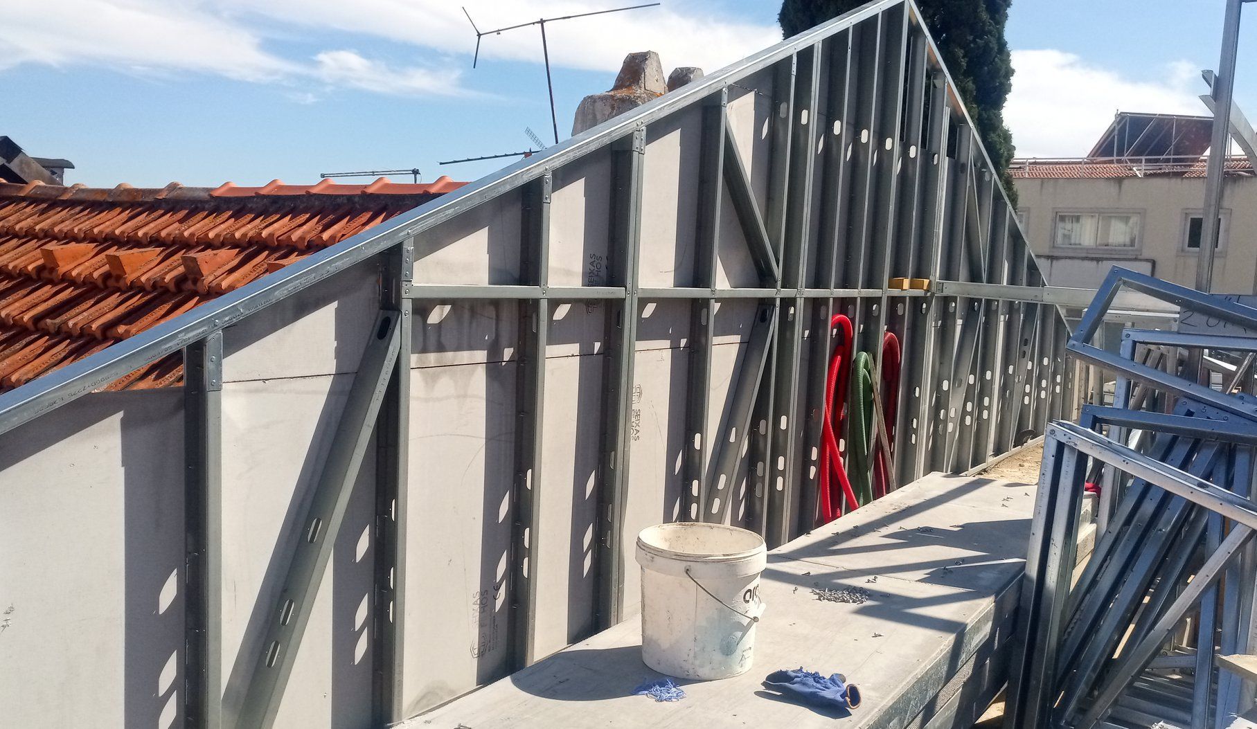 Estrutura metálica para o telhado de um edifício, com concreto e telhas vermelhas. Um balde de tinta está ao lado.