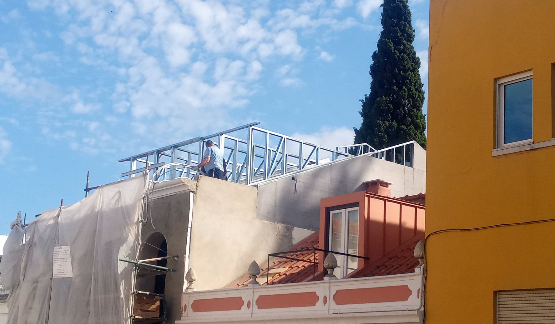 Canteiro de obras com estrutura metálica no telhado de um prédio, coberto por lonas brancas. Céu azul ao fundo.
