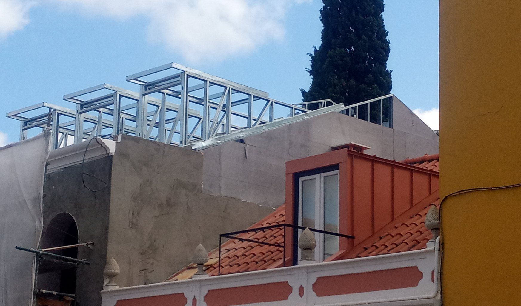Prédio em construção com estrutura metálica aparente no telhado. Uma árvore e céu azul ao fundo.