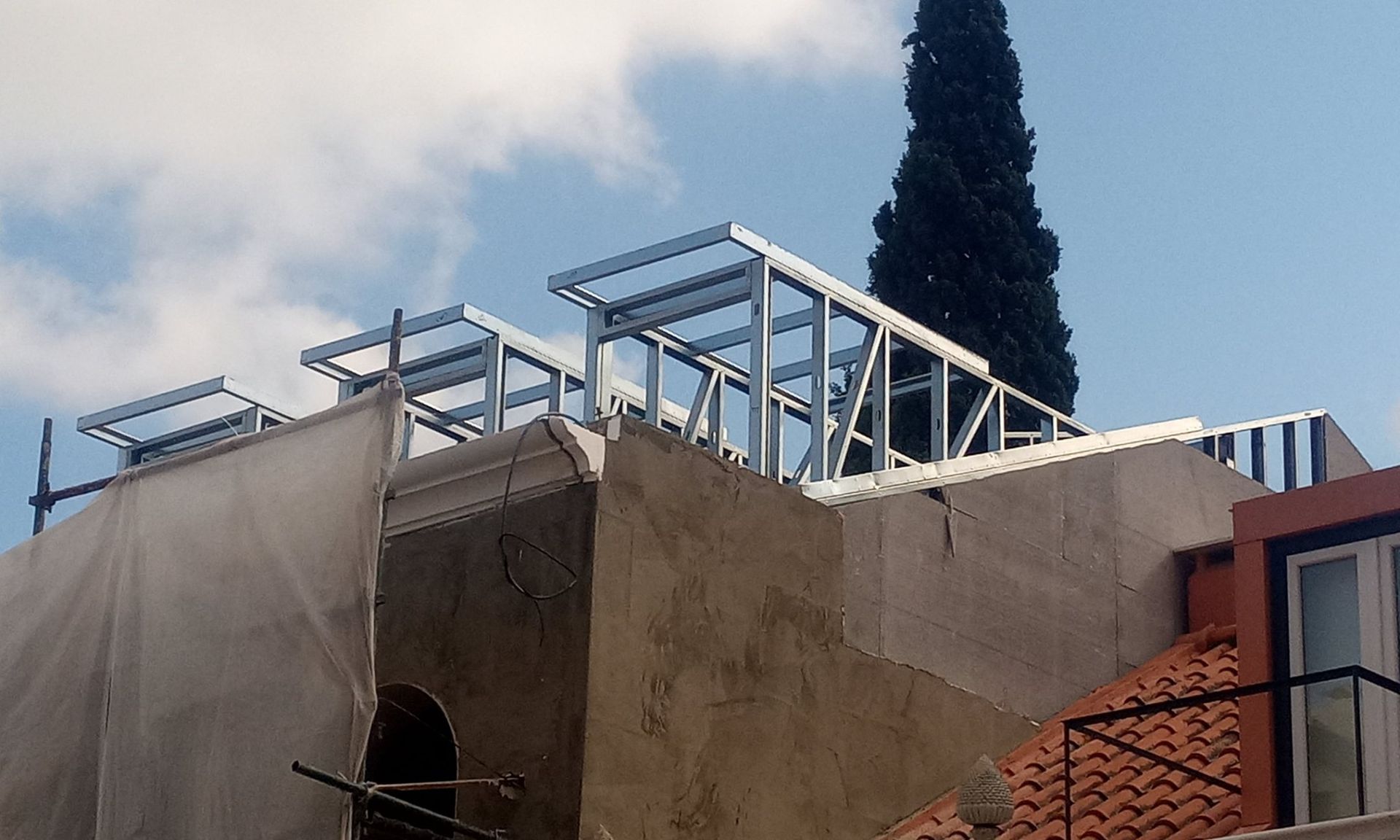 Estrutura metálica em construção no telhado de um edifício. Andaimes, árvores e parte do céu azul visíveis.
