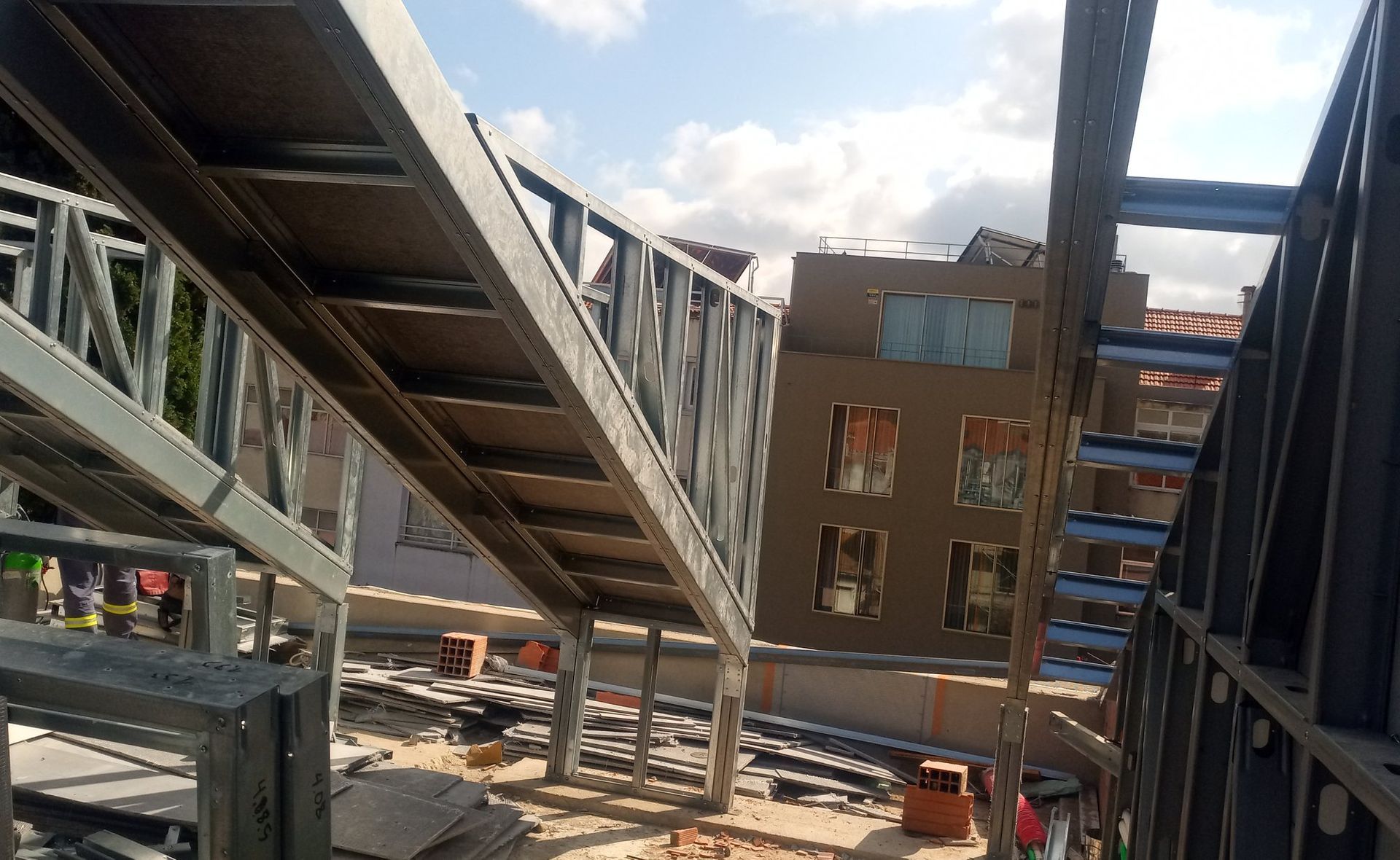 Estrutura metálica de um edifício em construção. Escada e estrutura em andamento, com um prédio ao fundo. Dia ensolarado.