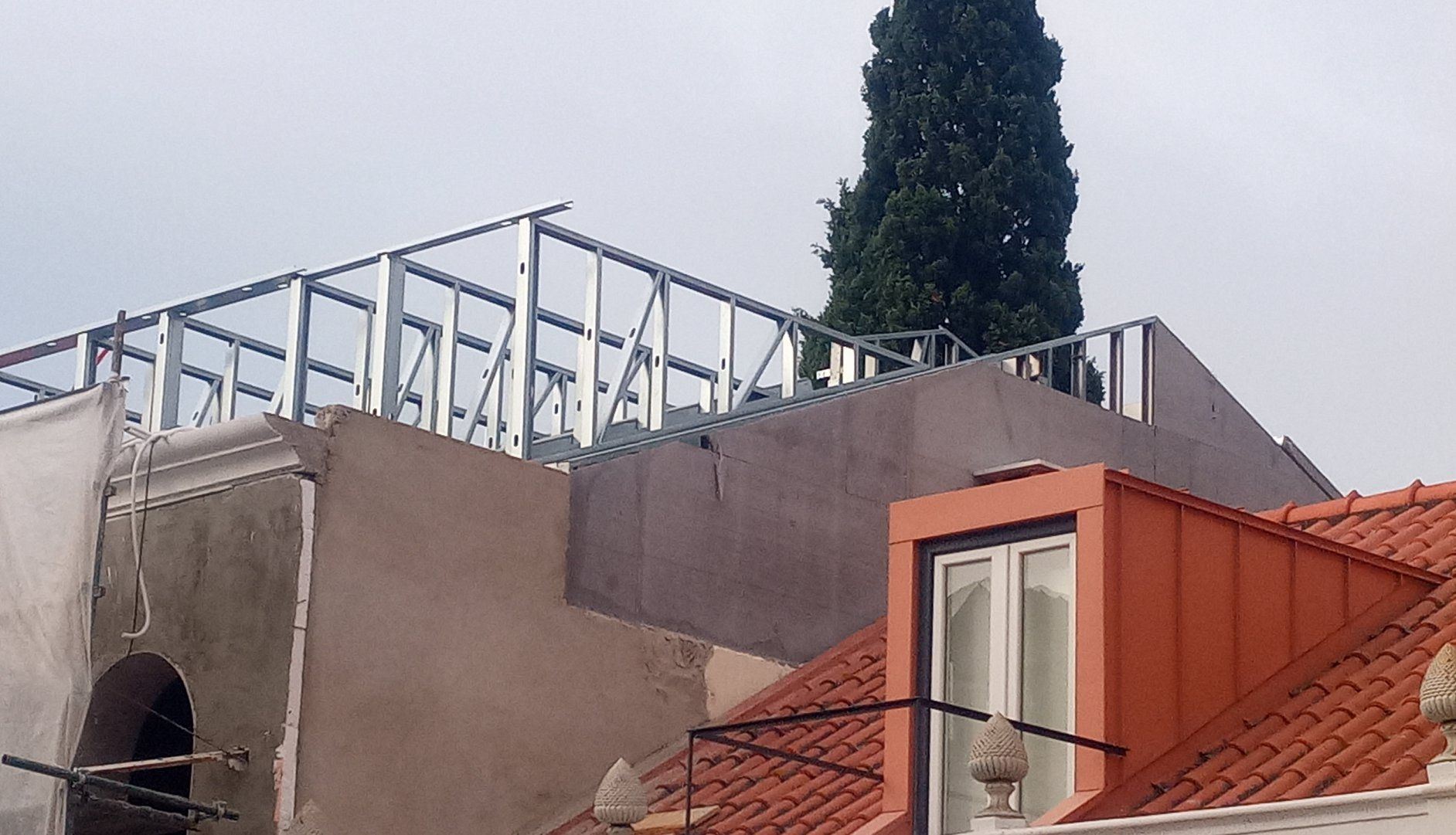 Construção de uma estrutura de cobertura com armação metálica, adjacente a um edifício com telhado de telha vermelha.