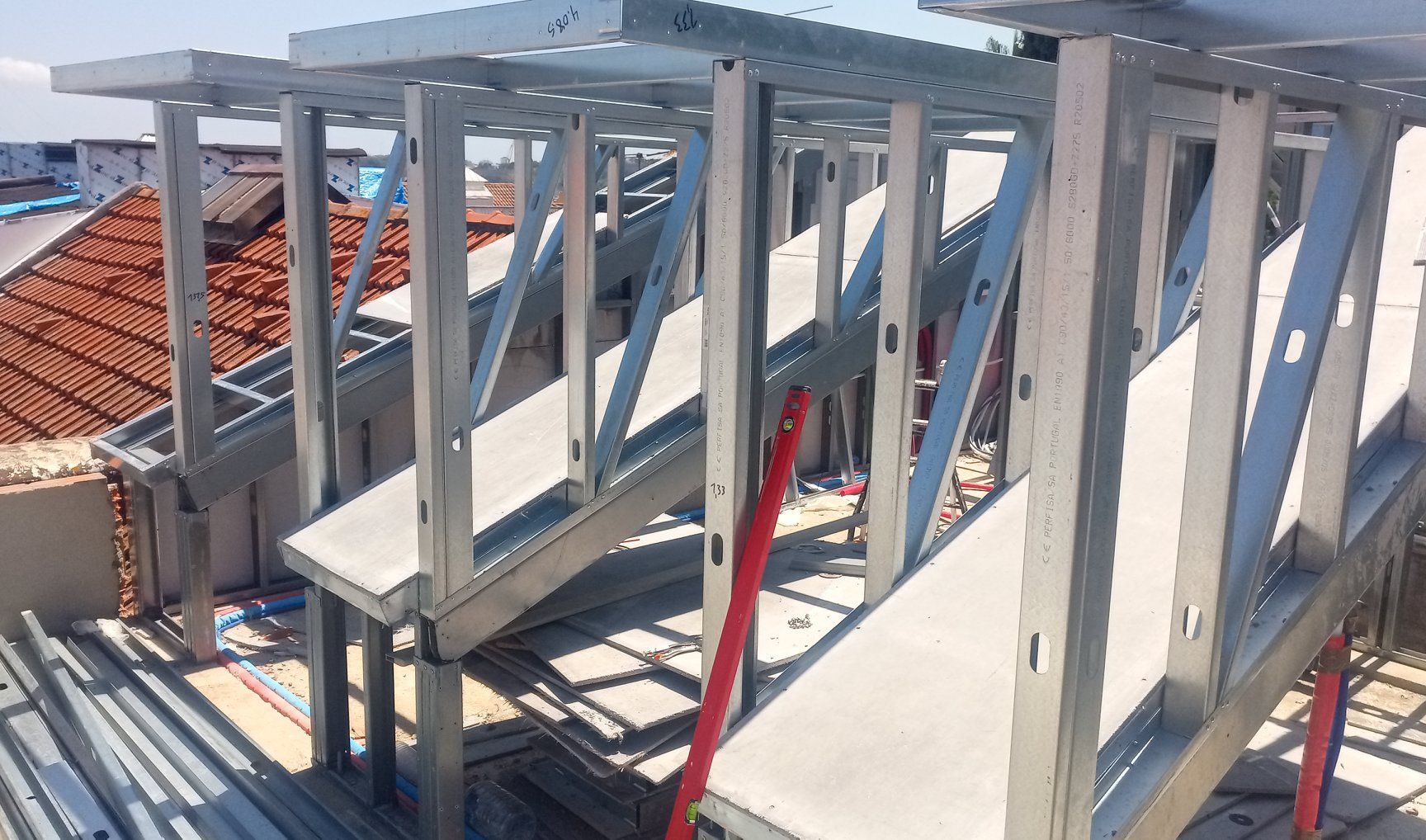 Estrutura metálica em um telhado, possivelmente para construção. Vigas cinzentas com um cabo vermelho. Prédio de tijolos ao fundo.