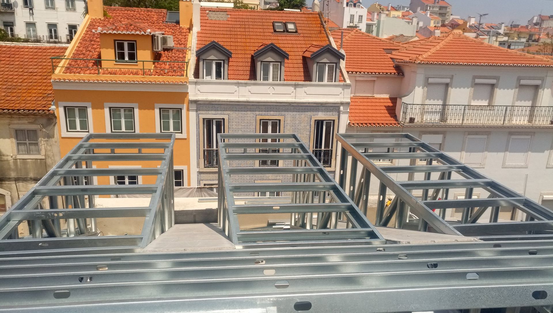 Estrutura metálica do telhado de um edifício, com prédios laranja e brancos ao fundo.
