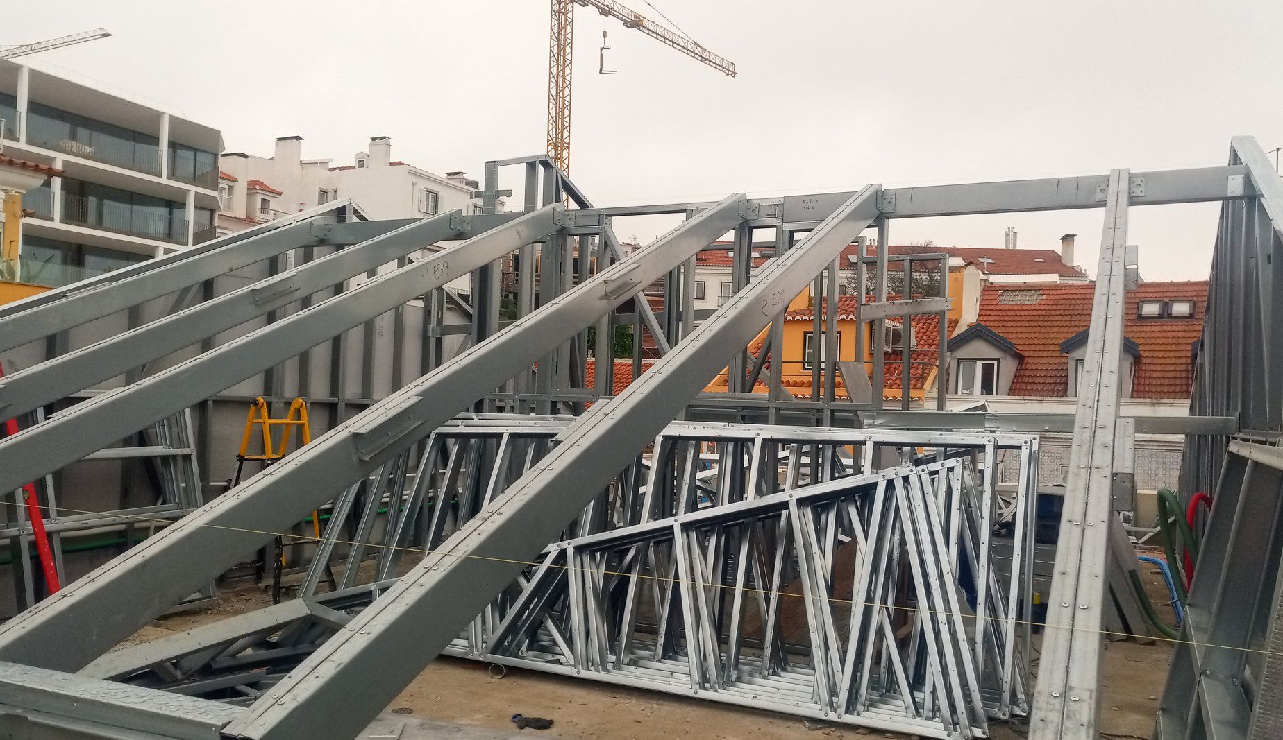 Canteiro de obras com estrutura metálica, guindaste e edifícios circundantes.
