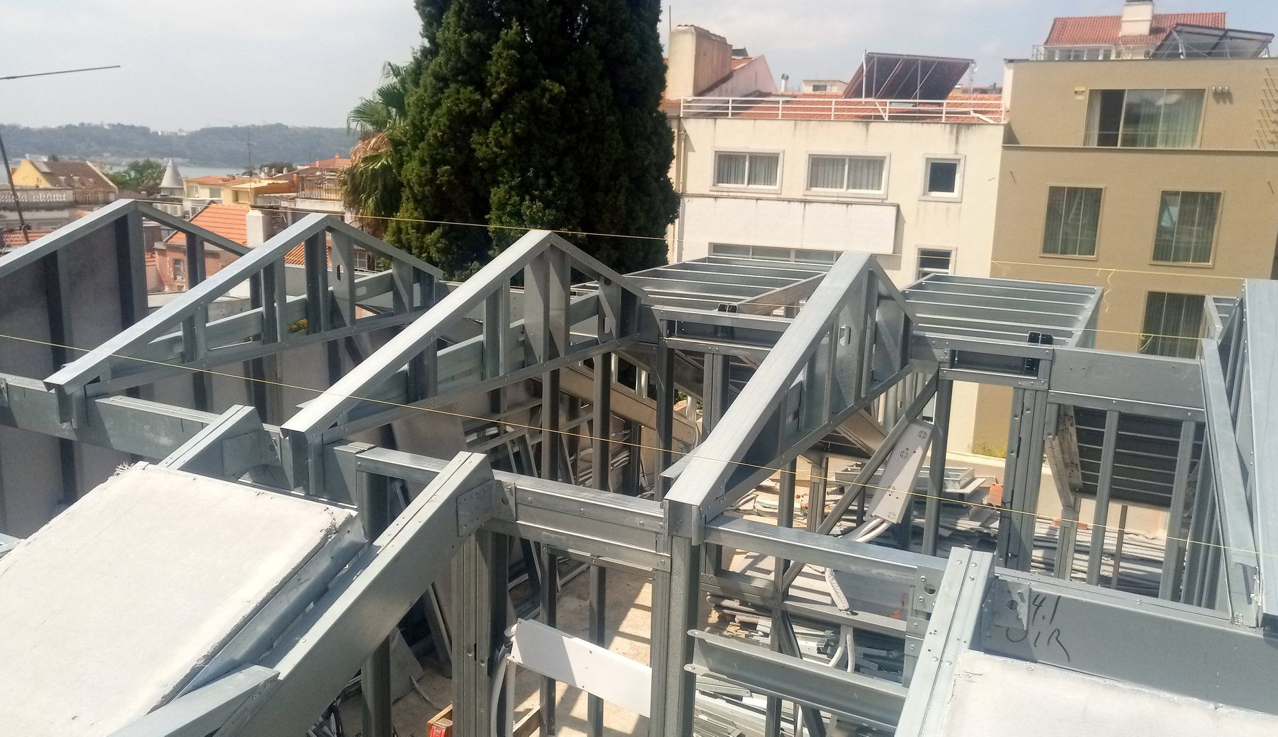 Estrutura de aço de um edifício em construção sobre um telhado, com prédios da cidade e uma árvore ao fundo.