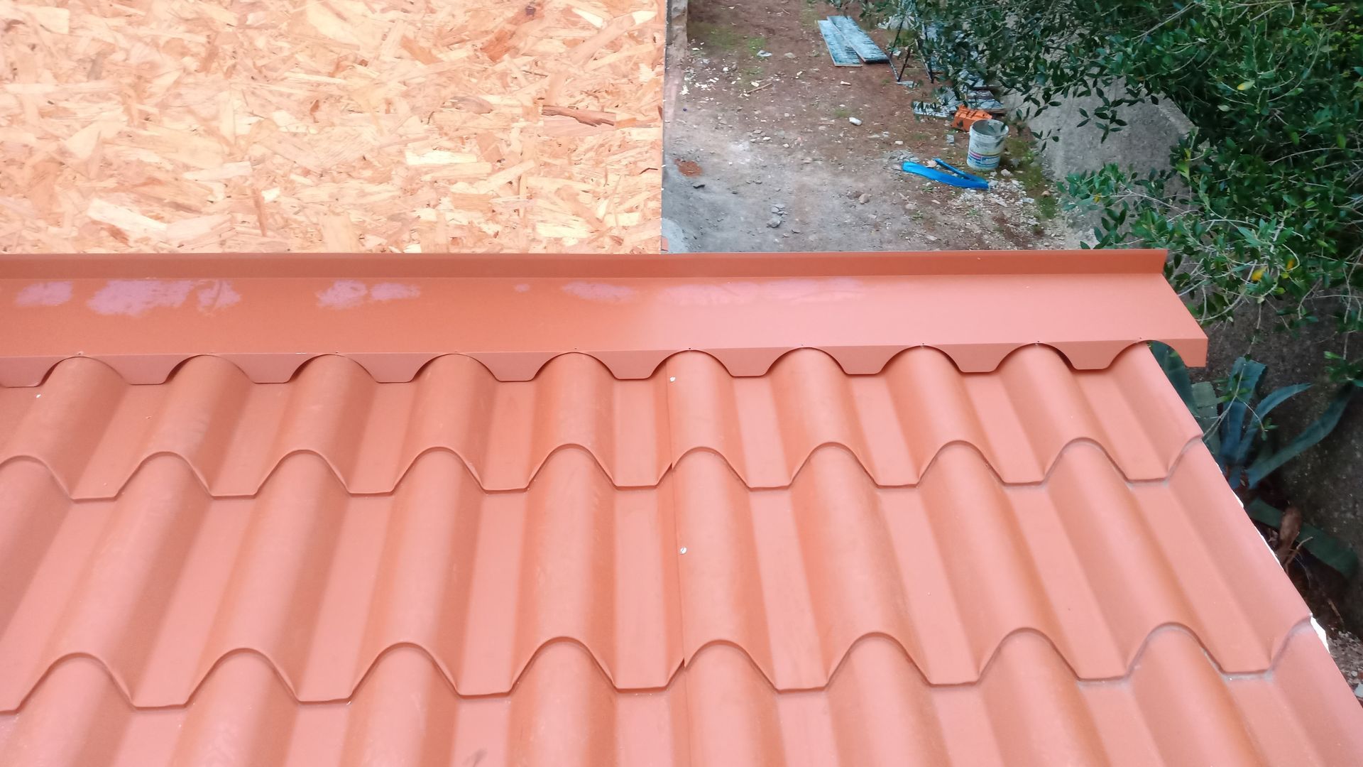 Telhado de telha vermelha com borda metálica combinando, contra uma parede marrom-clara, com superfície de concreto sujo e vegetação ao fundo.