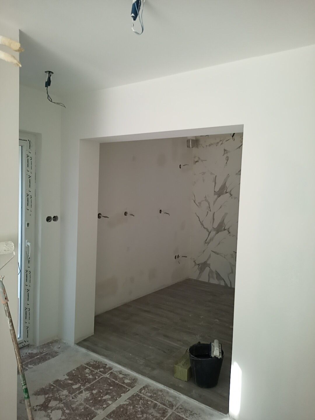 Porta interna que dá acesso a um pequeno cômodo inacabado com fiação exposta e piso de concreto. Paredes brancas, balde à vista.