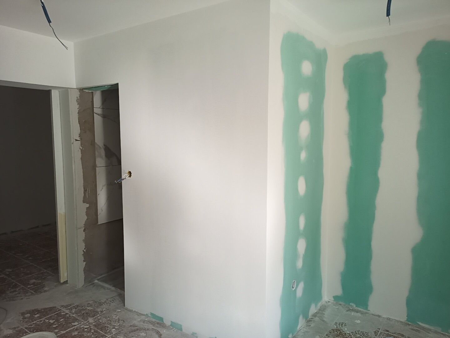 Interior de um cômodo com paredes brancas, parte da parede de gesso acartonado verde coberta e uma porta aberta.
