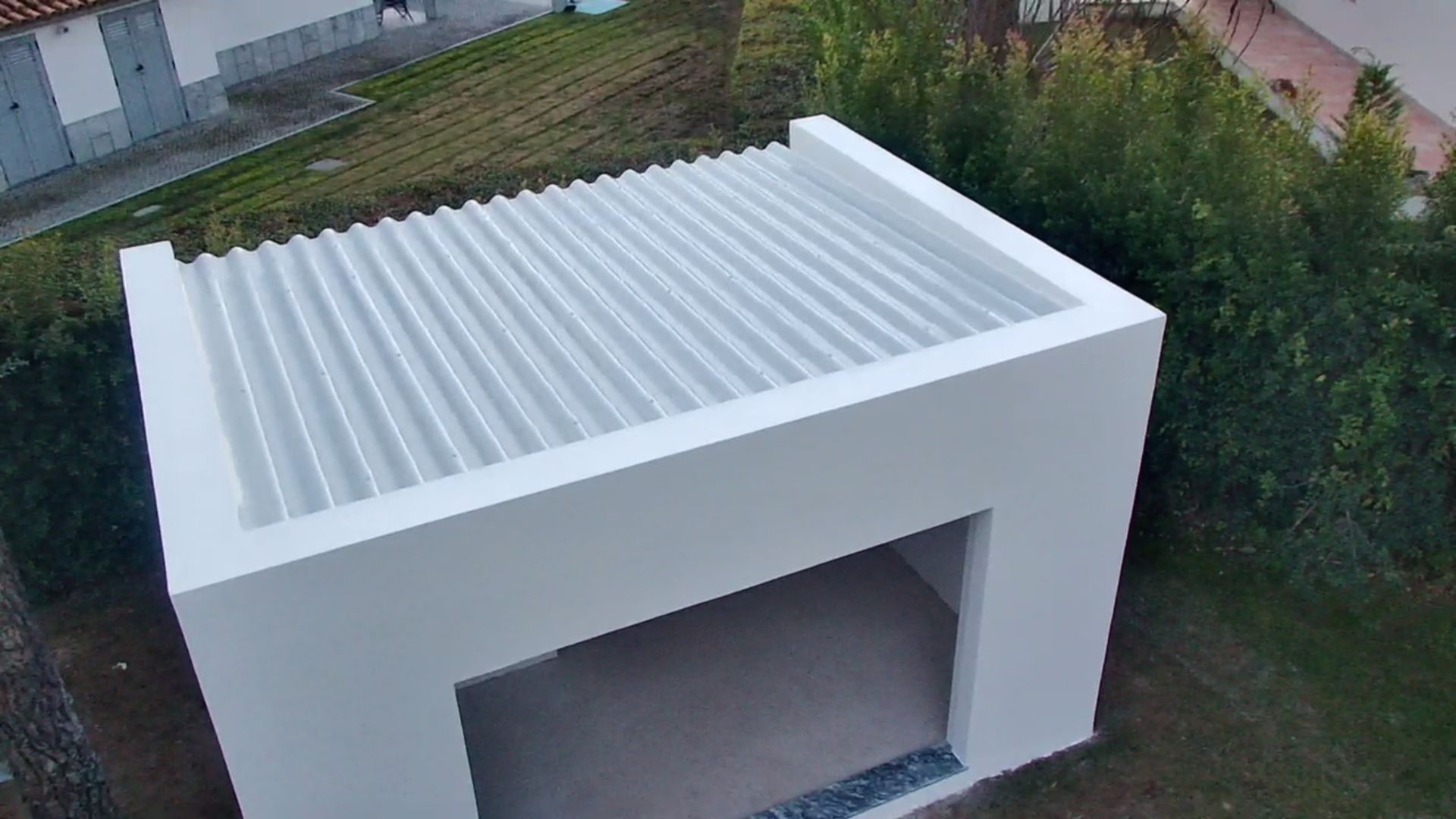 Prédio branco, em formato de cubo, com telhado de metal ondulado e fachada aberta; cercado por folhagem verde.