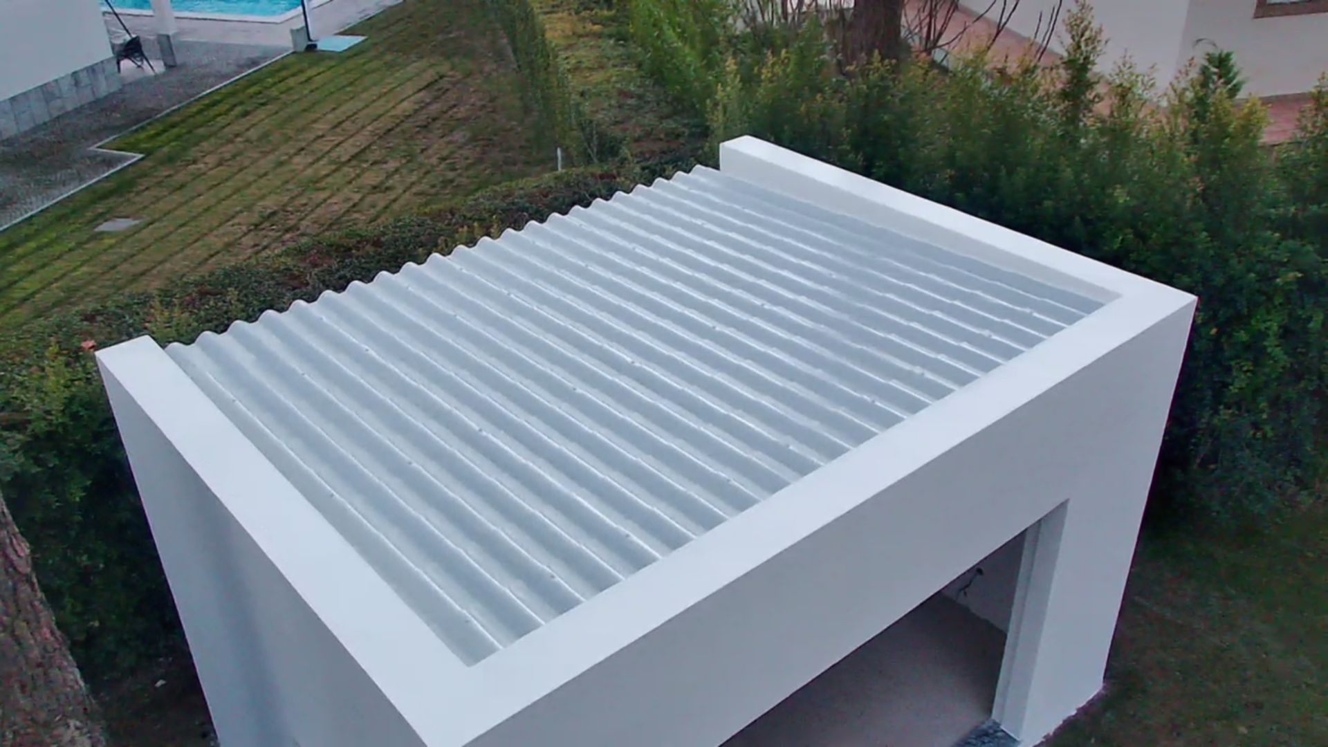 Estrutura externa branca e moderna com telhado ondulado, vista de cima, rodeada de vegetação.