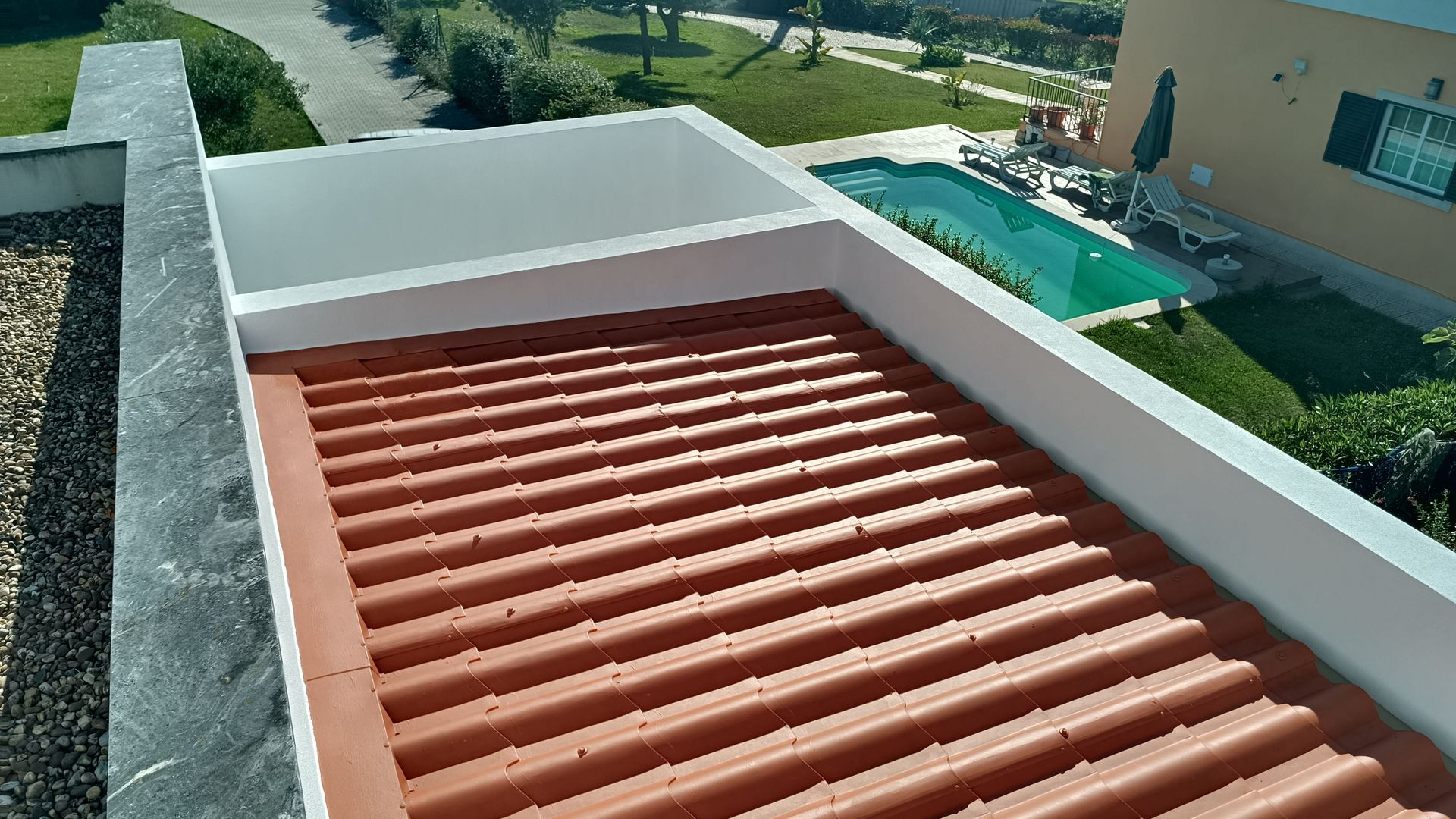 Telhado de telha de terracota com paredes brancas, com vista para uma piscina e jardim num dia ensolarado.