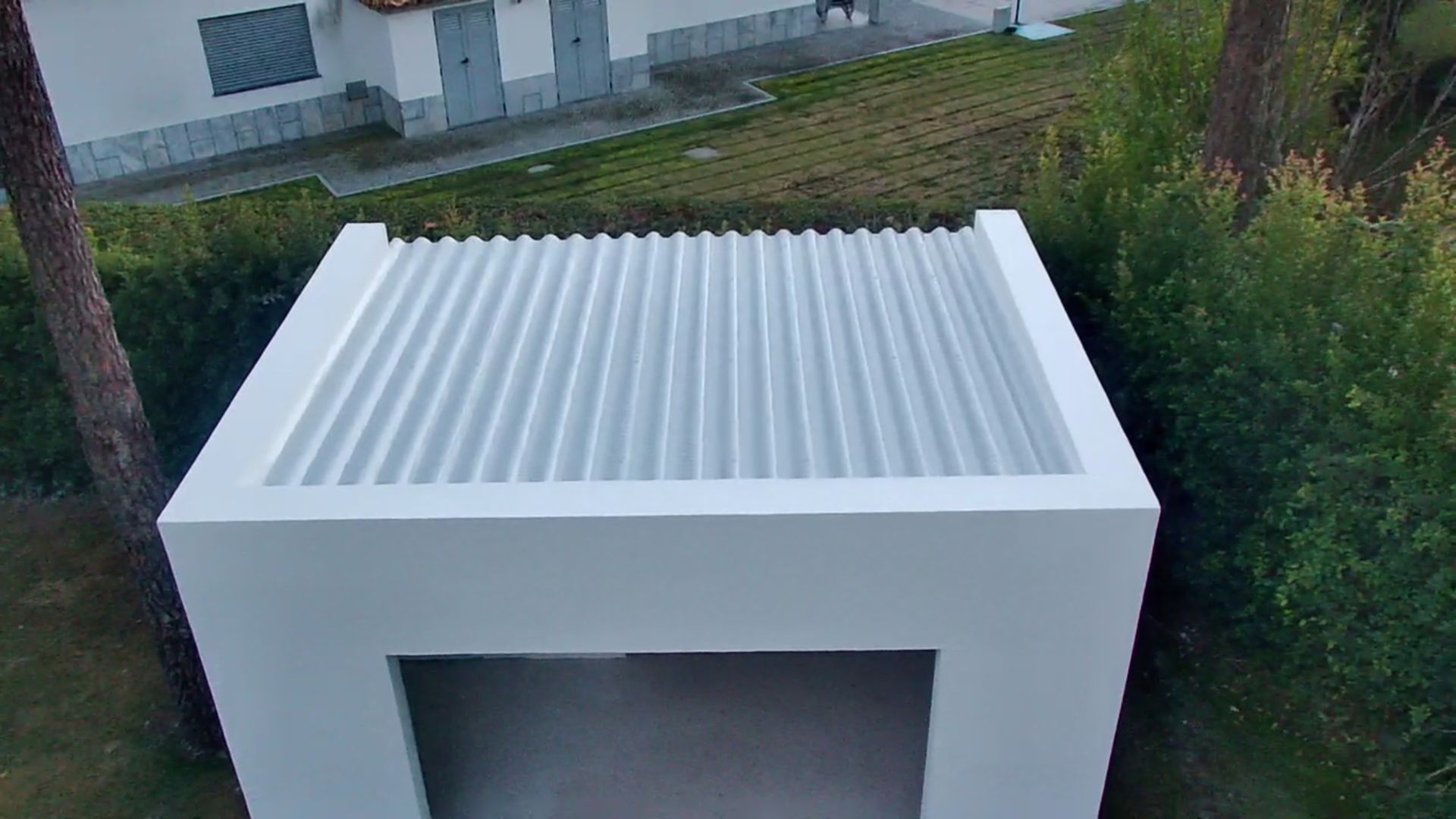 Estrutura branca em forma de caixa com telhado de zinco. Está localizada em uma área gramada perto de árvores e um prédio.