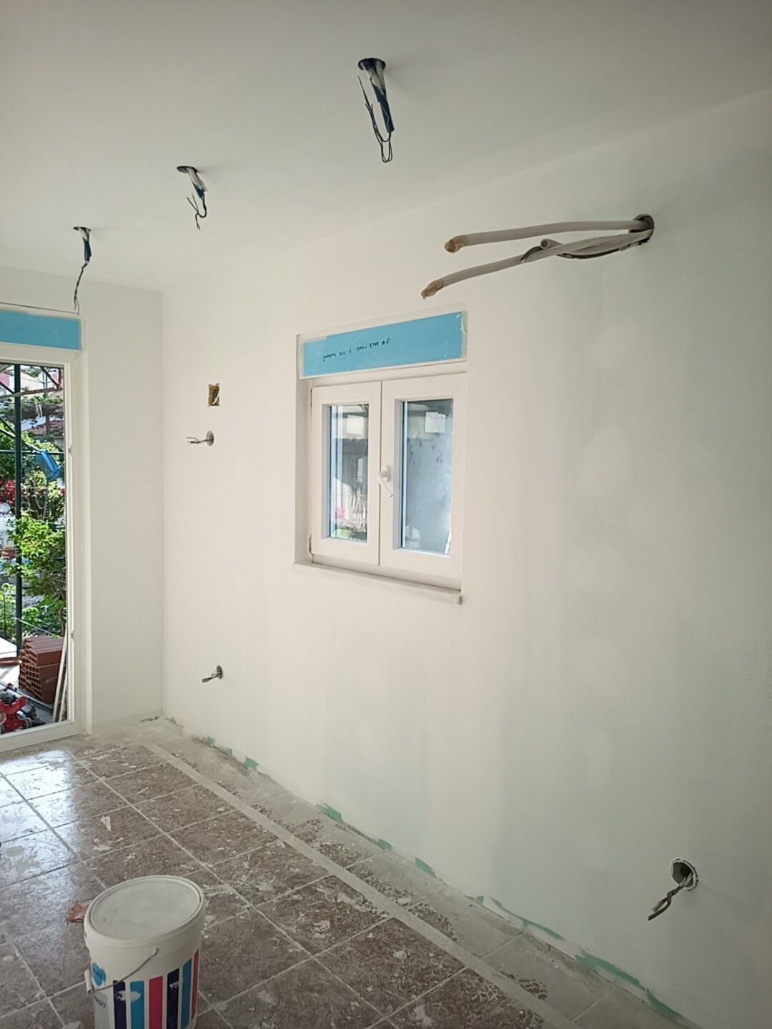 Interior de um cômodo em reforma com fiação exposta, uma janela e um balde de tinta.