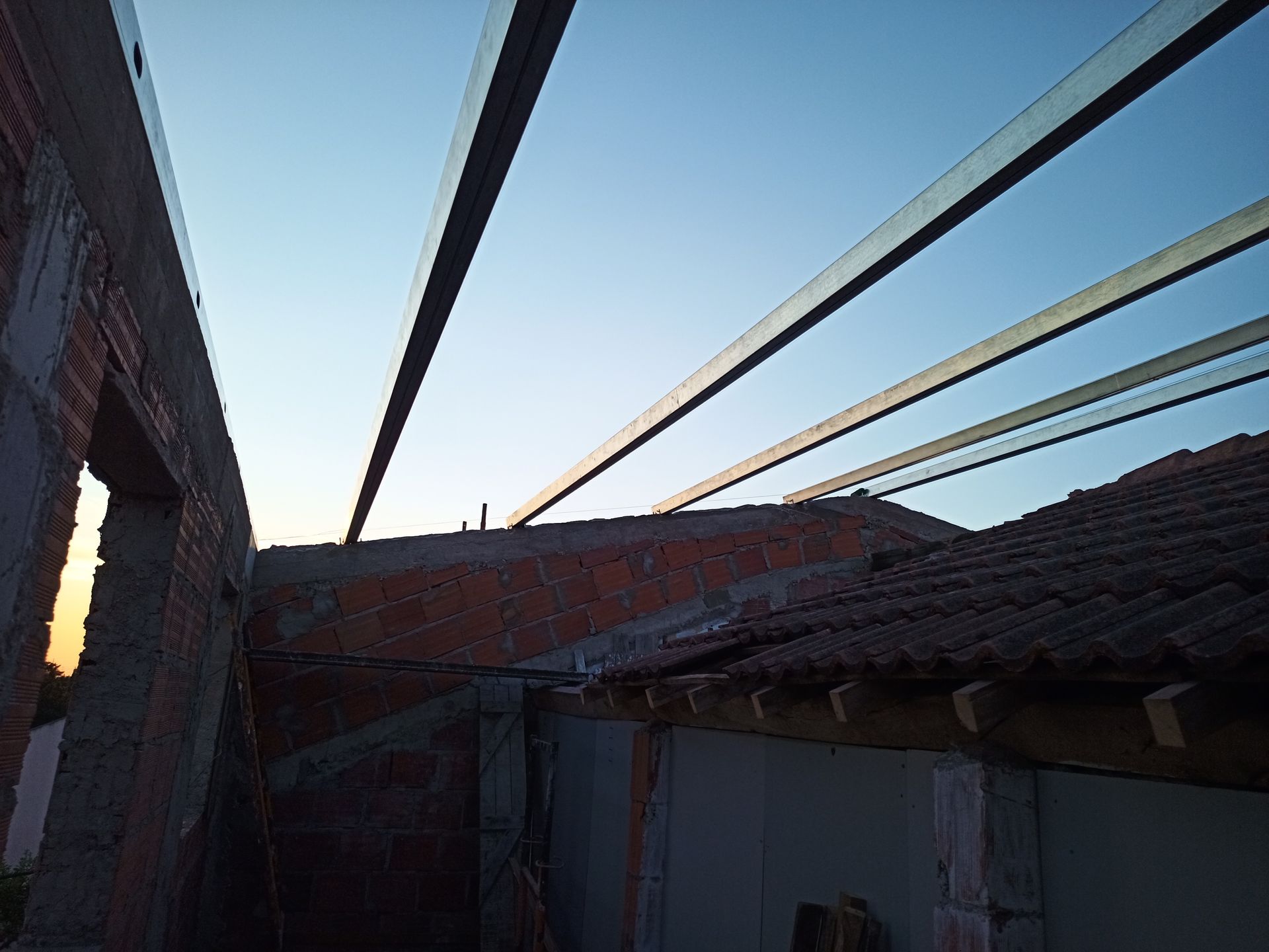 Vigas de aço que se estendem de uma parede de tijolos em direção ao telhado, contra um céu azul ao pôr do sol.