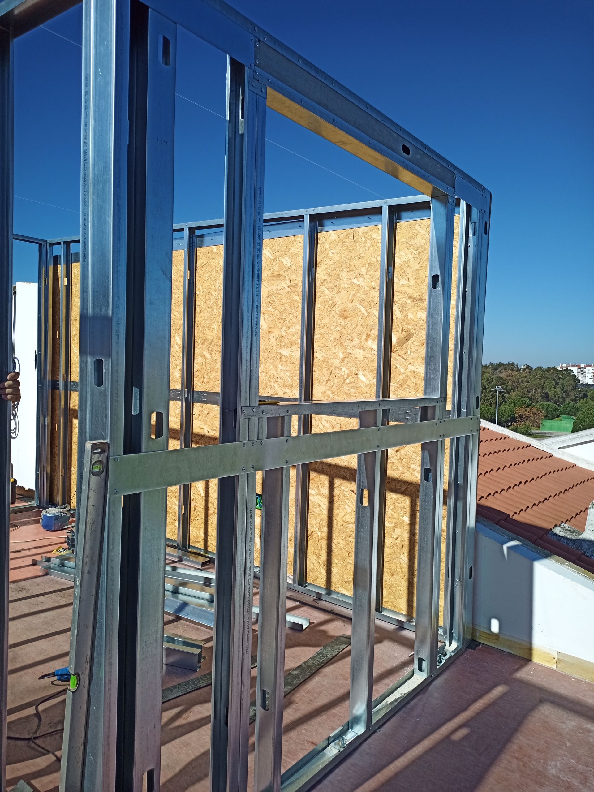 Estrutura metálica de um edifício em construção, com painéis de madeira no interior da estrutura, contra um céu azul.