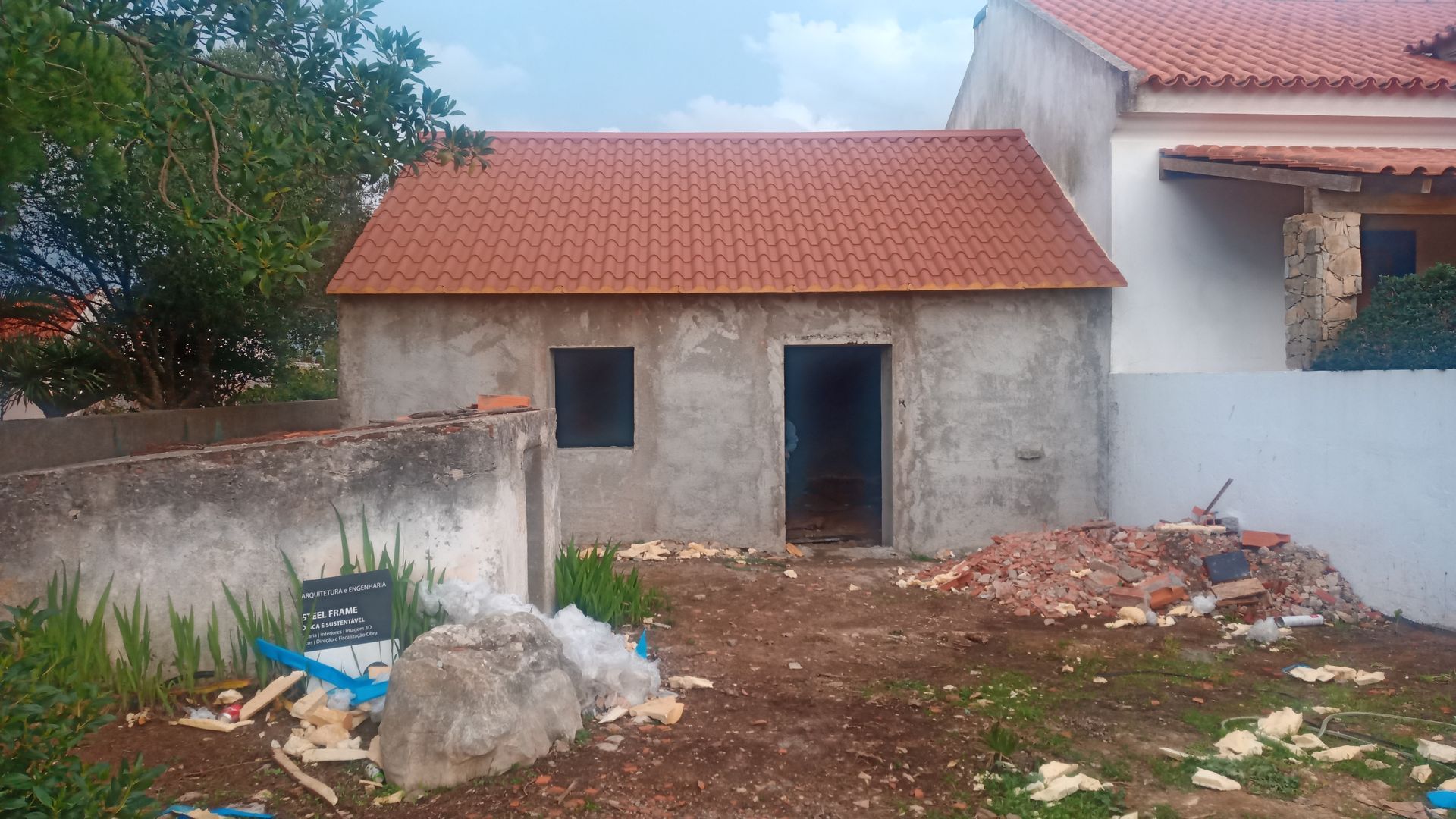 Pequeno prédio de concreto inacabado com telhado de telhas vermelhas; entulho de construção no quintal.