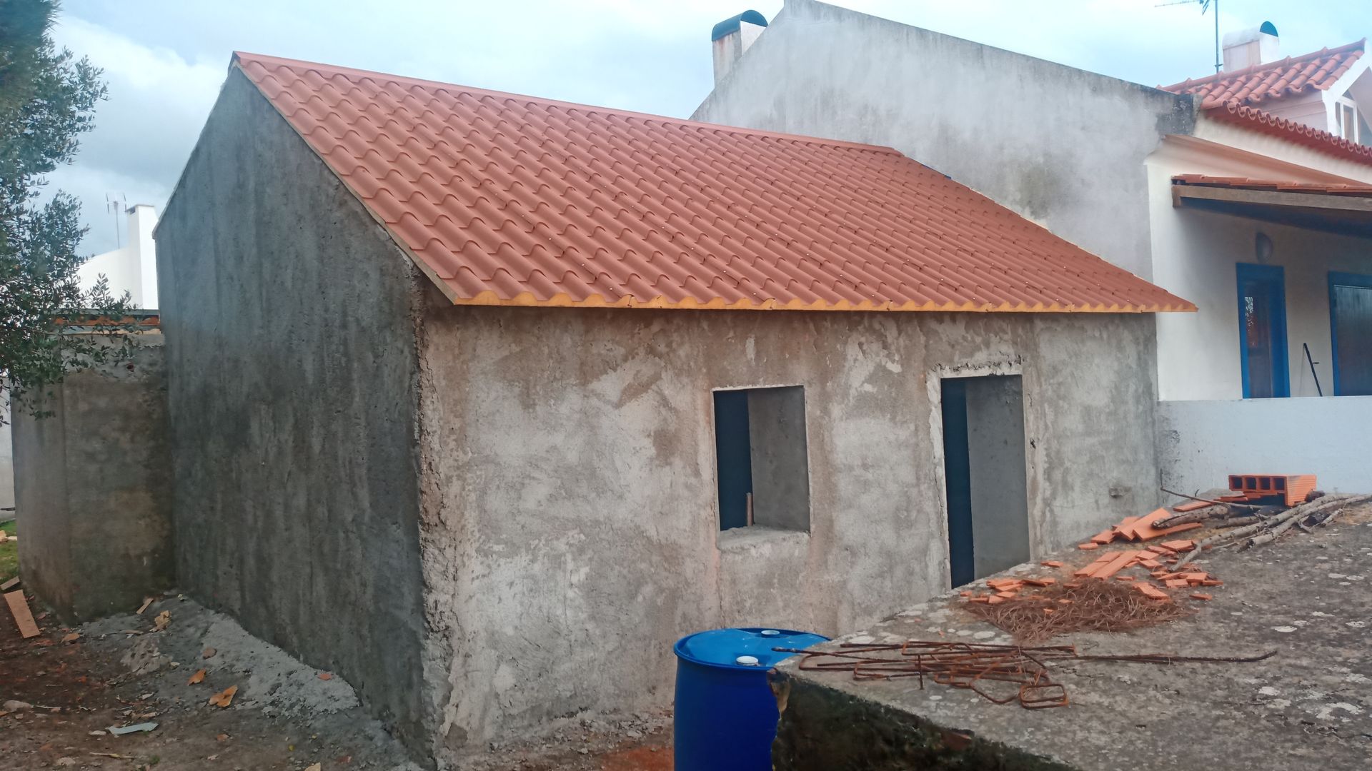 Pequeno prédio inacabado de concreto com telhado de telha vermelha, janela e batente de porta. Construção externa.
