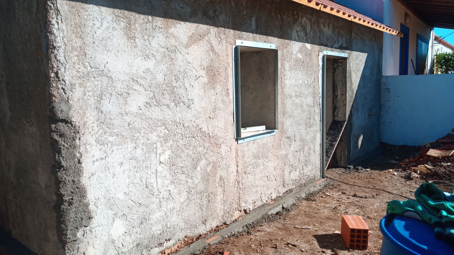 Vista exterior de um pequeno edifício em construção, paredes de estuque cinza, duas janelas e entulho ao redor da base.