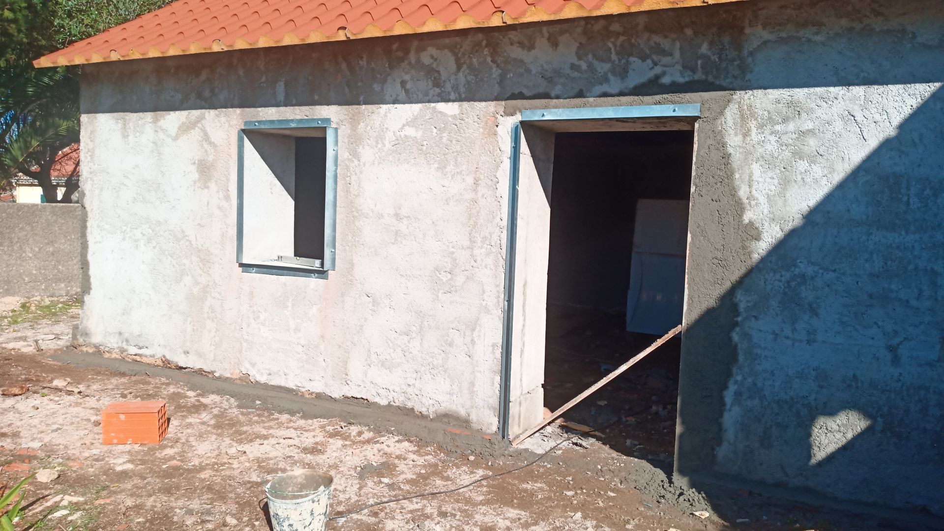 Vista exterior de um pequeno edifício em construção; paredes de estuque, telhado laranja, porta aberta e uma pequena janela com moldura azul.