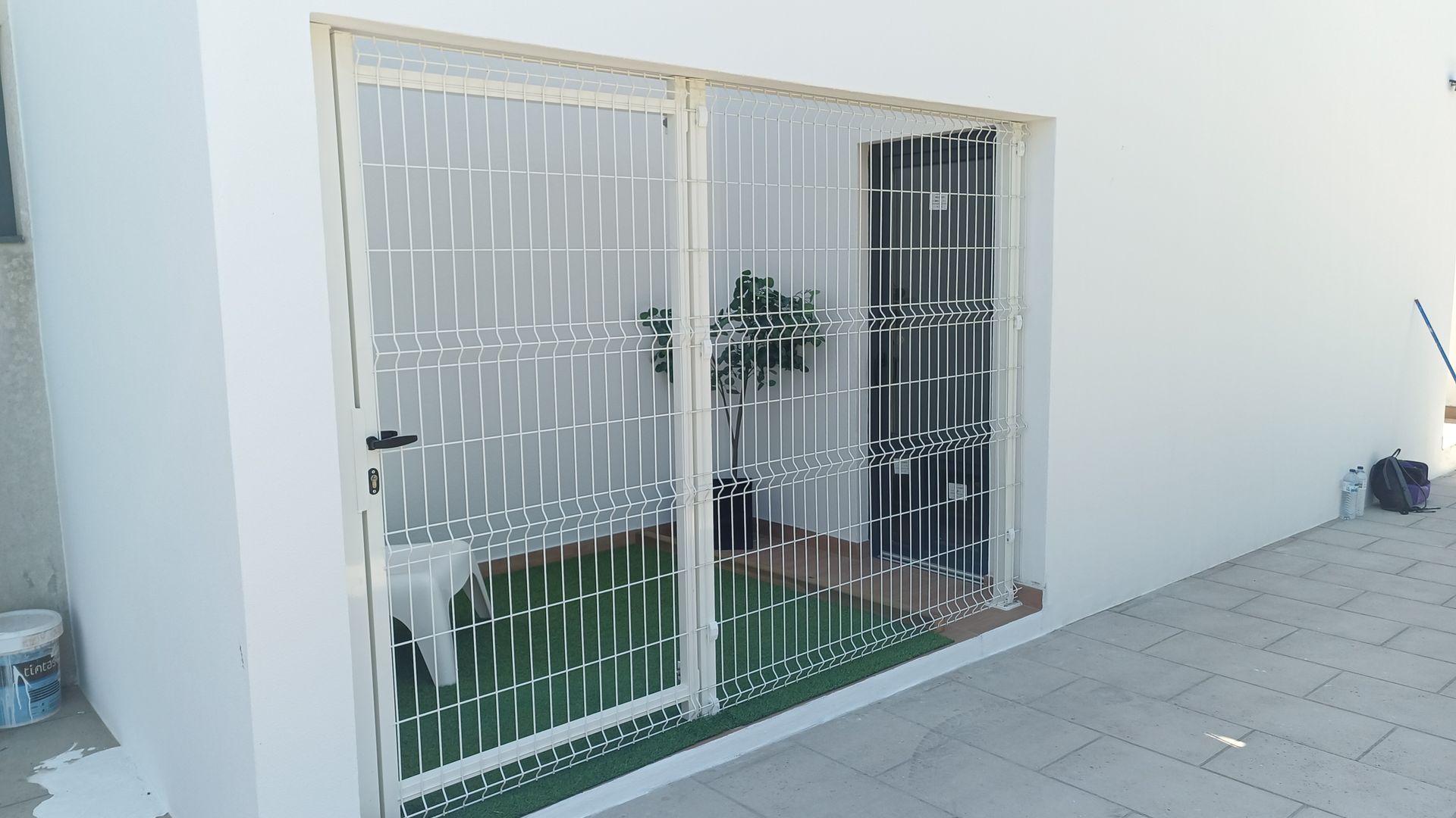 Um prédio branco com portões de segurança sobre a entrada, contendo grama artificial e um vaso de planta.