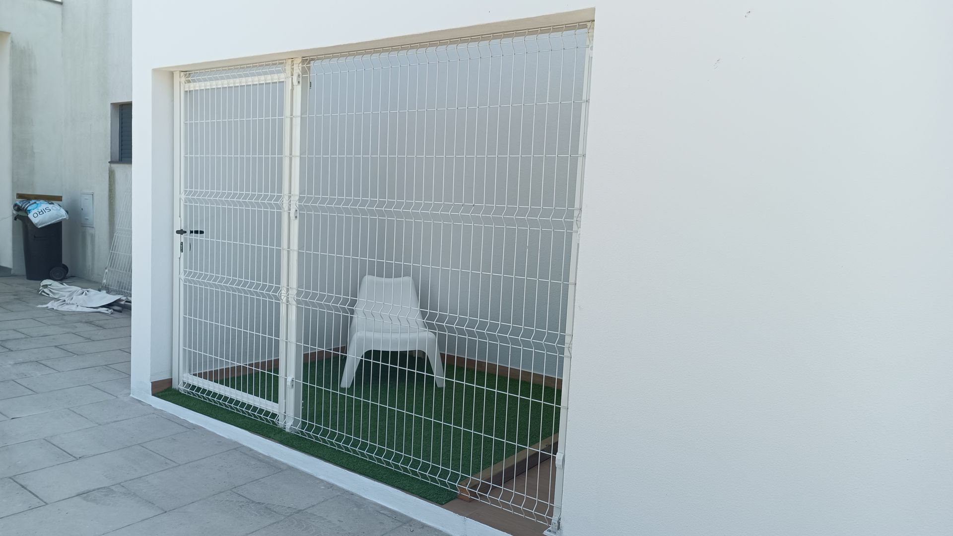 Portão de segurança branco em frente a um pequeno pátio com grama verde e uma cadeira branca.