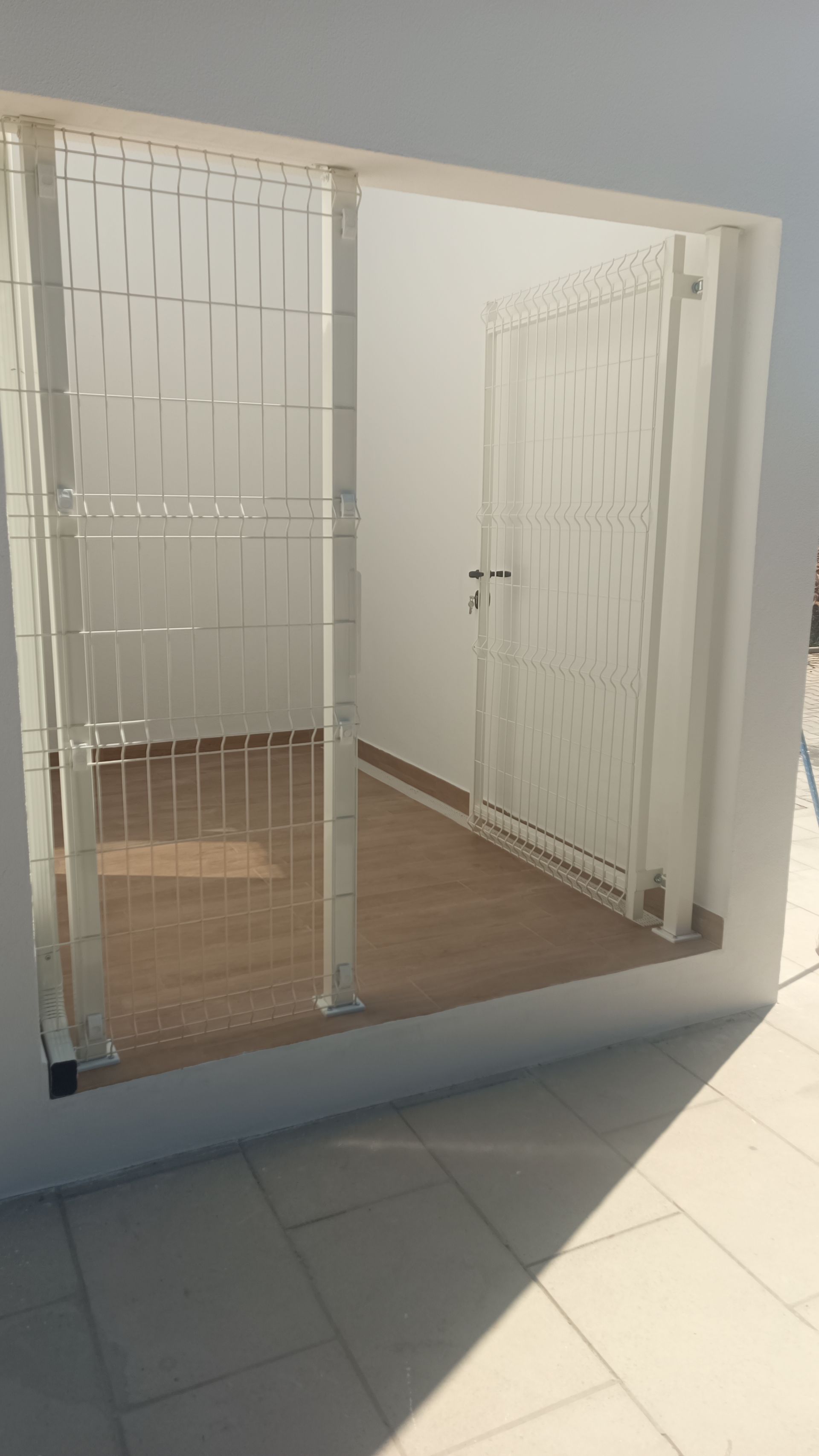 Um portão e uma porta de segurança brancos delimitam uma pequena sala vazia com piso que imita madeira.