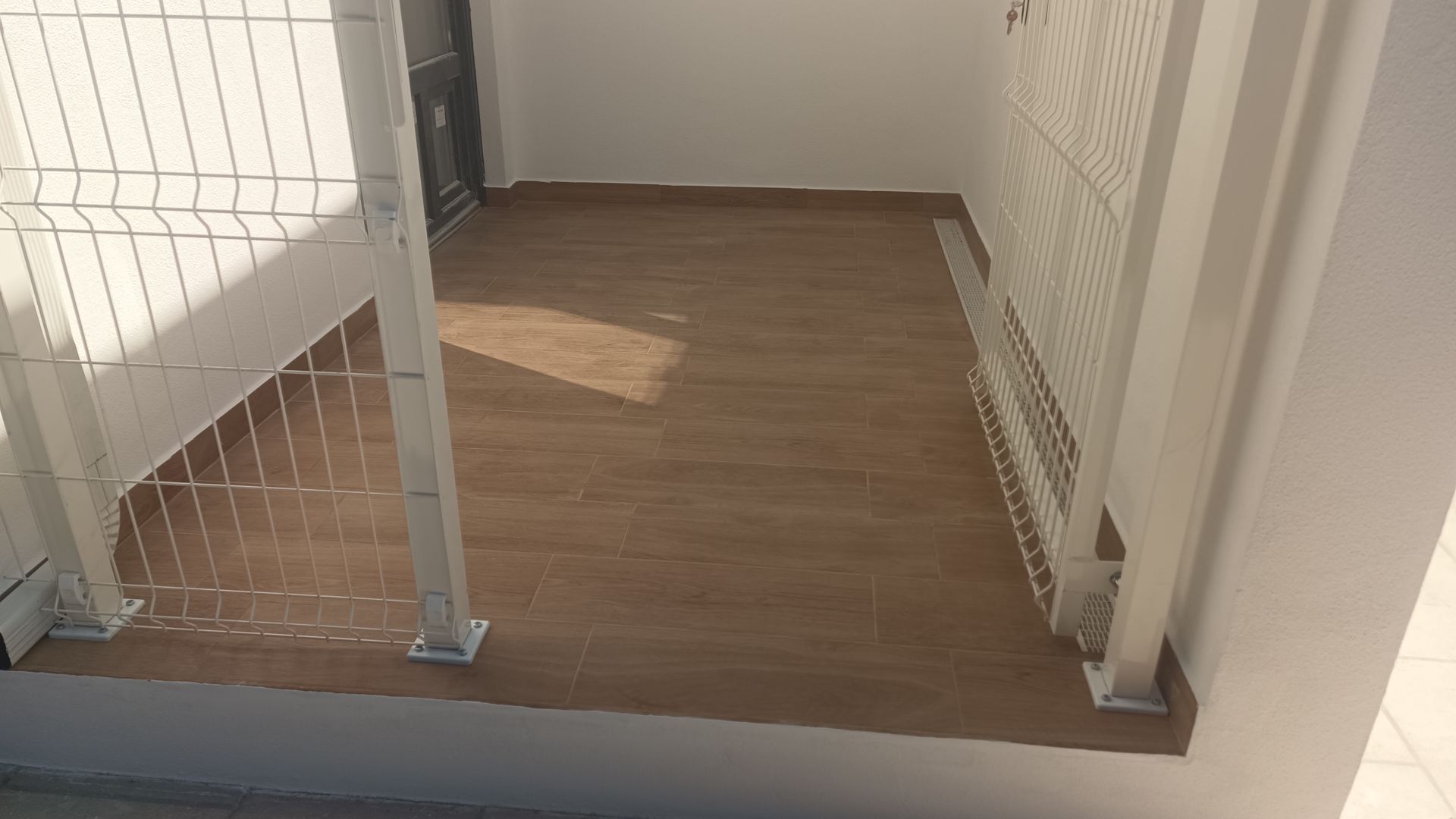 Uma pequena varanda com piso imitando madeira e grades de metal branco, ao lado de uma parede branca.