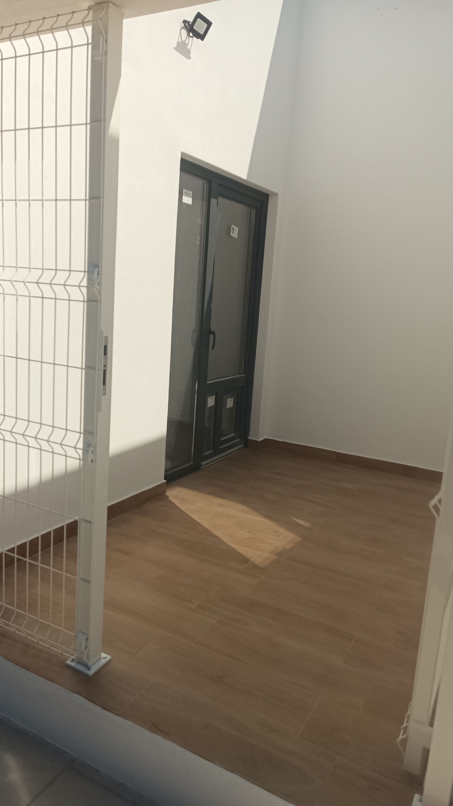 Vista interior: porta de canto, piso de madeira, paredes brancas e luminária.