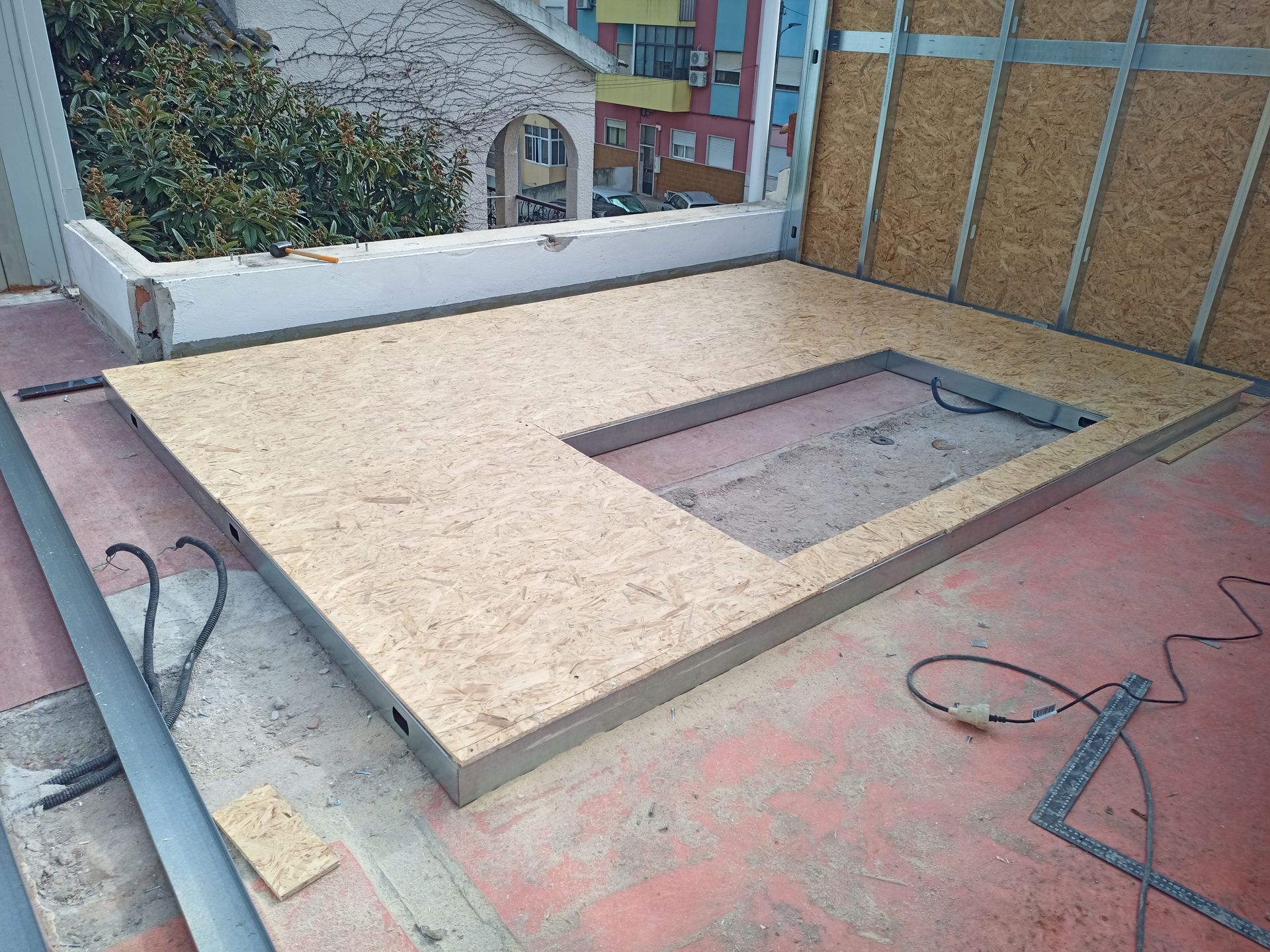 Canteiro de obras: Painel de piso OSB com abertura retangular assentado sobre concreto vermelho. Estrutura metálica visível.