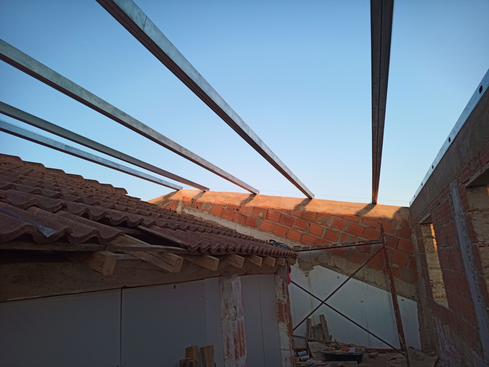 Vigas metálicas estendendo-se sobre uma parede de tijolos e um telhado de telhas, com um céu azul claro ao fundo.