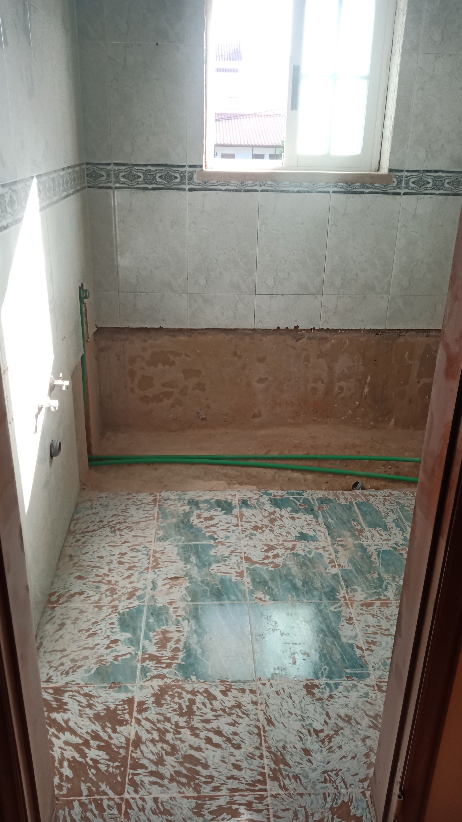 Reforma do banheiro em andamento; parede danificada, canos expostos e piso parcialmente azulejado.