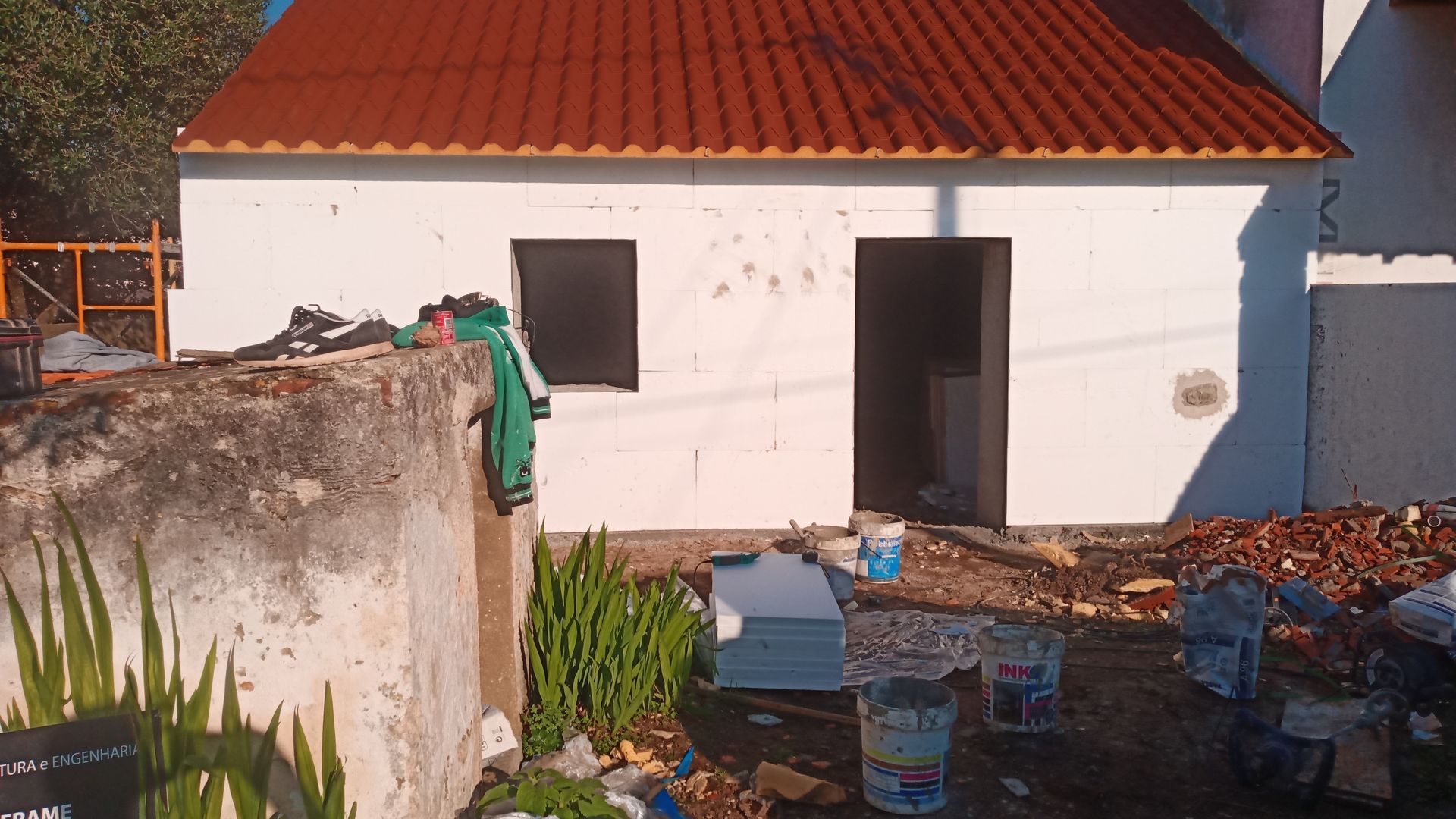Pequeno prédio branco em construção; telhado laranja, porta e janela pretas, ferramentas e entulho espalhados do lado de fora.