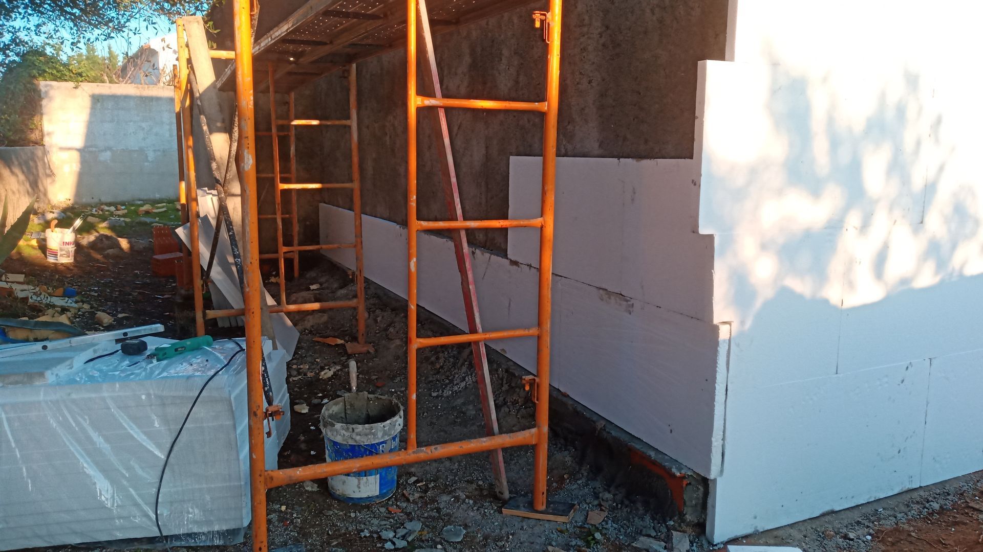 Cena de construção: andaimes laranja, painéis de isolamento branco fixados na parede e baldes no chão.