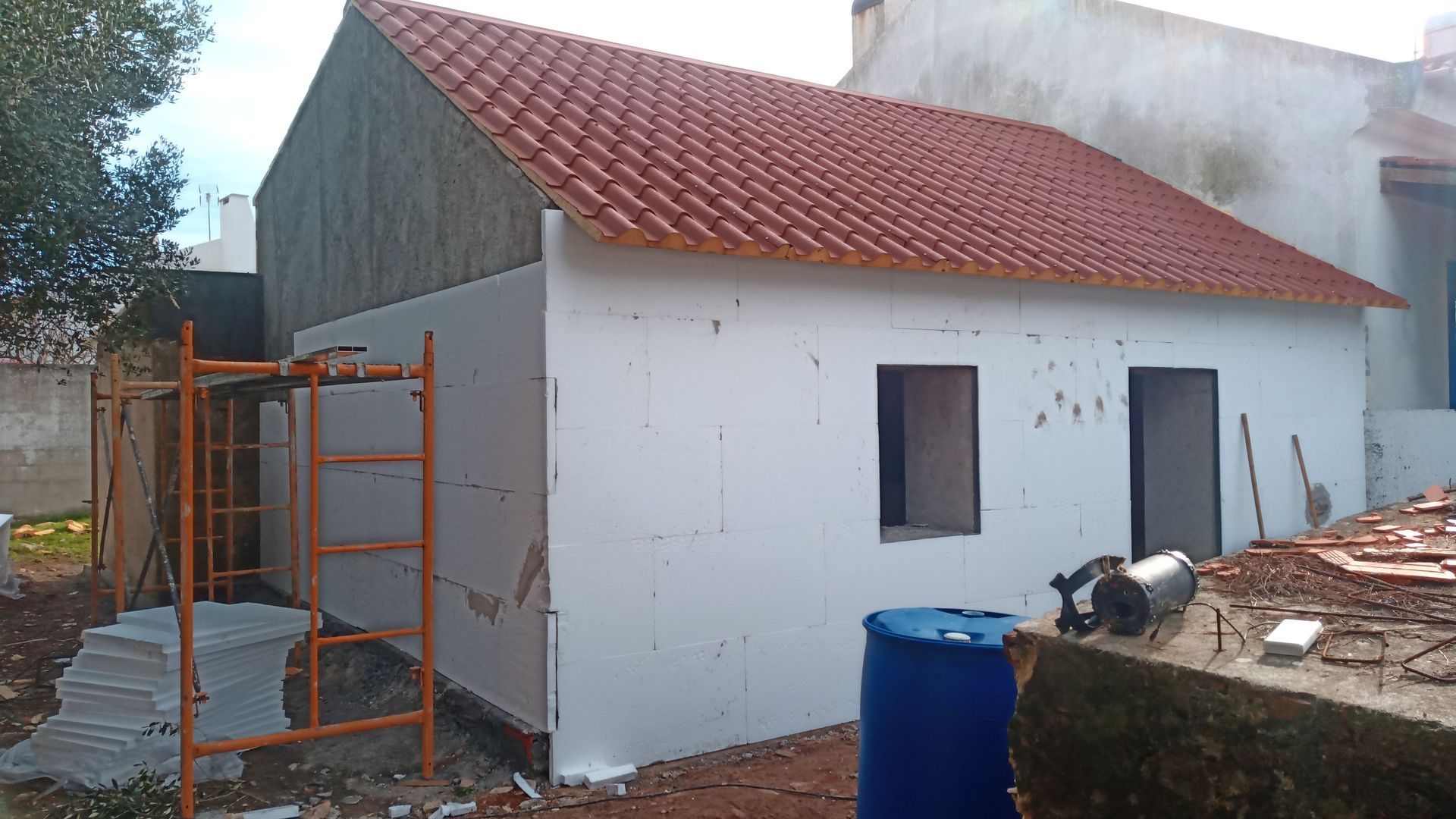 Pequena casa branca em construção, com telhado de telhas laranja, andaimes e barril azul.