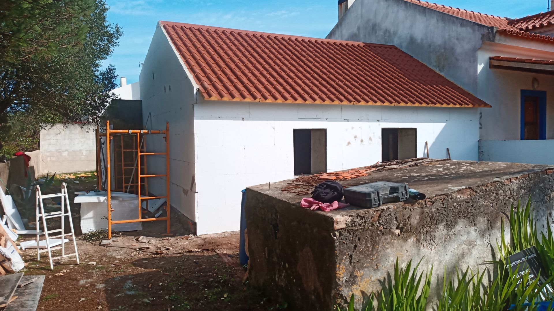 Prédio branco em construção com telhado de telha vermelha, andaimes e estrutura de pedra em primeiro plano.