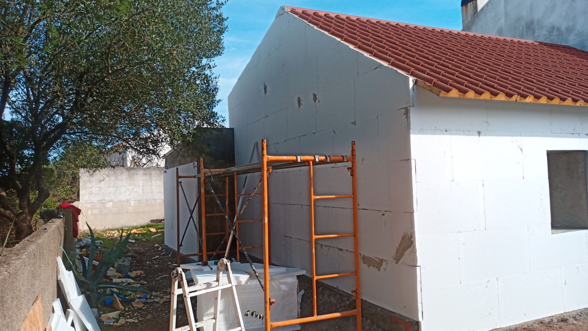 Casa em construção; painéis de isolamento branco nas paredes externas, andaimes laranja, telhado de telha vermelha.