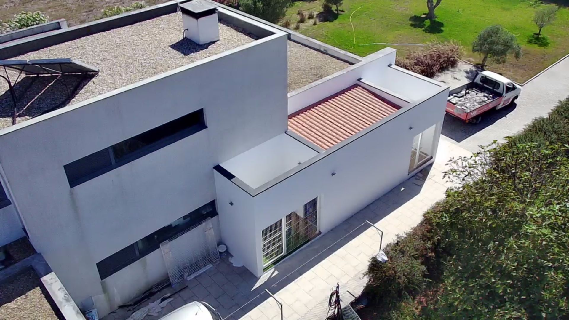 Casa moderna branca com telhado plano, painéis solares e uma seção do telhado com telhas vermelhas. Um caminhão está estacionado nas proximidades.