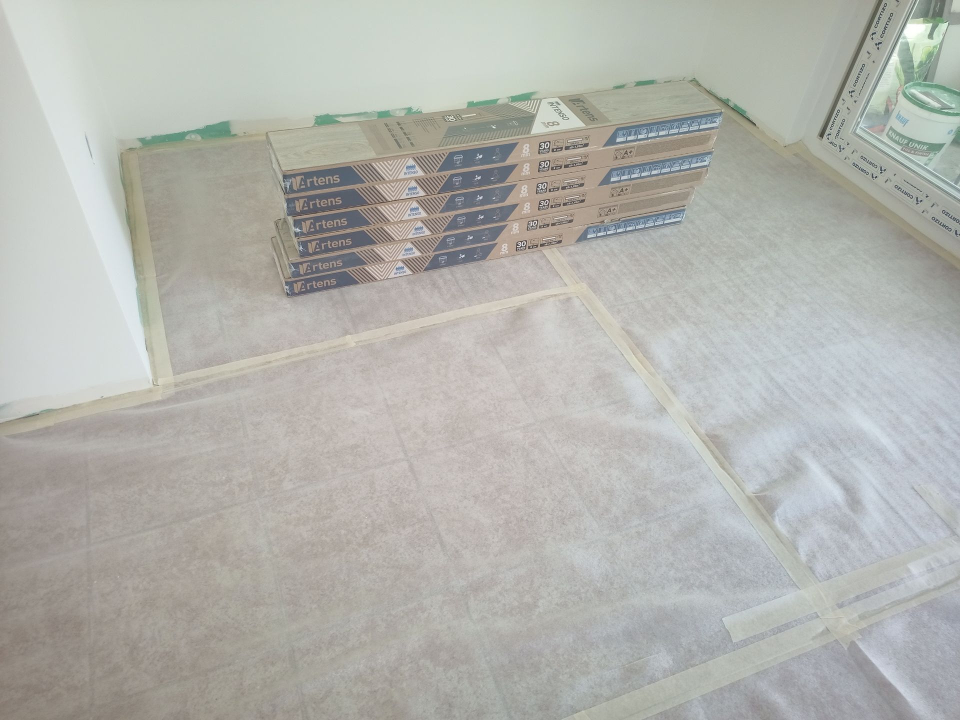 Pilha de ladrilhos de piso sobre papel protetor em um cômodo, perto de uma janela.