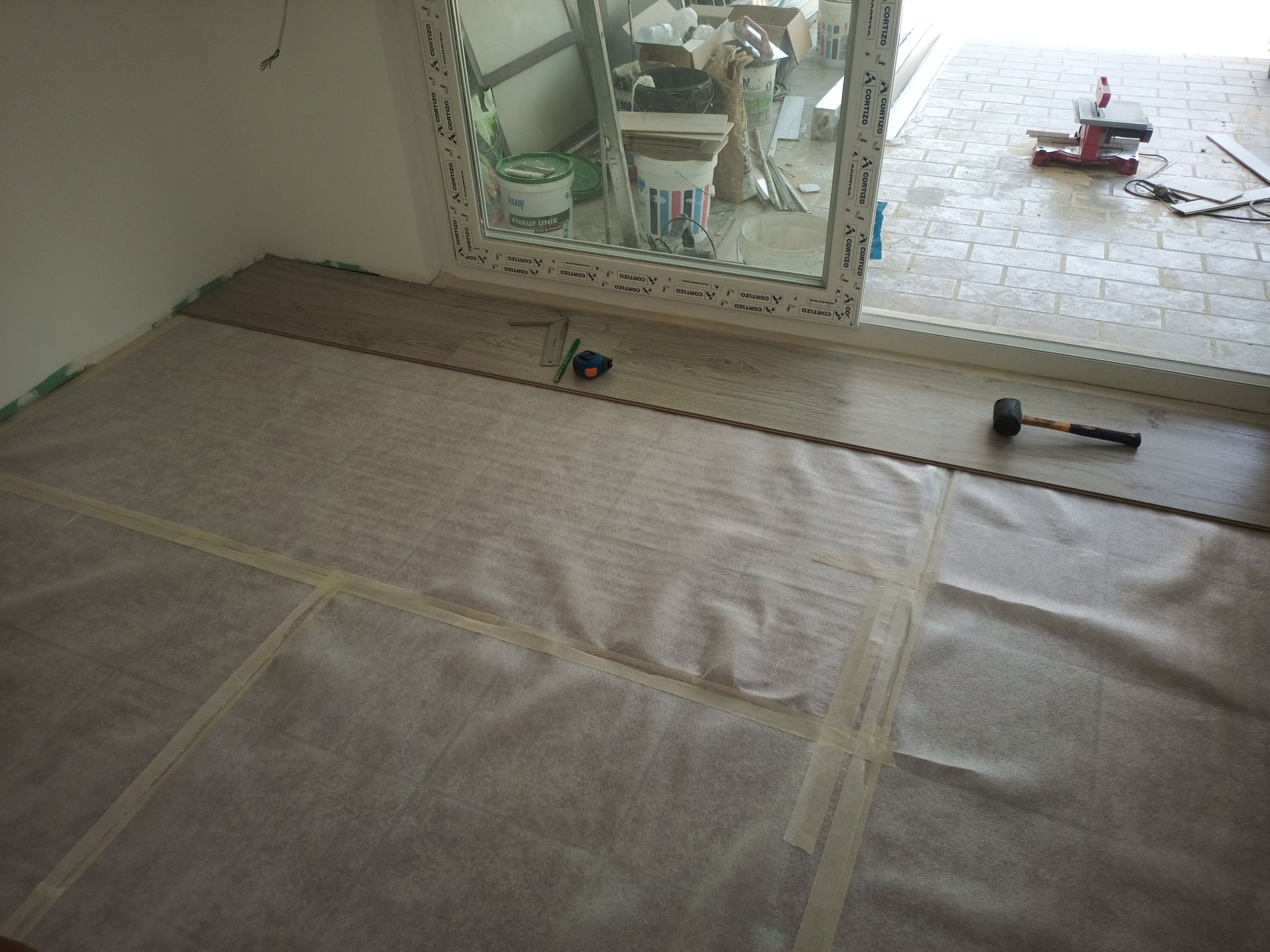 Instalação do piso em andamento; tábuas de madeira assentadas sobre uma base branca ao lado de uma janela.