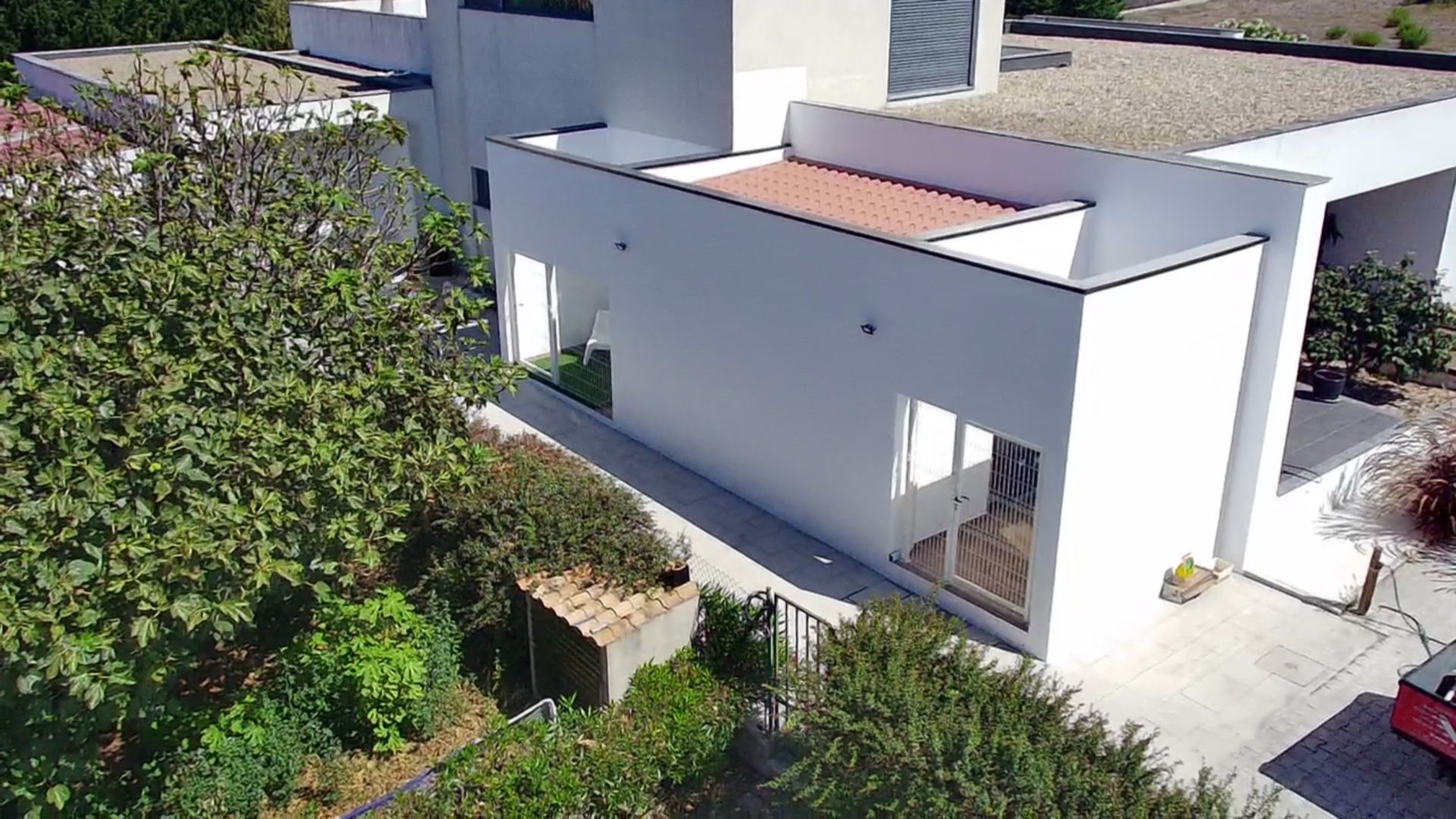 Prédio moderno branco com telhado plano e pátio com telhas vermelhas, rodeado por folhagem verde.