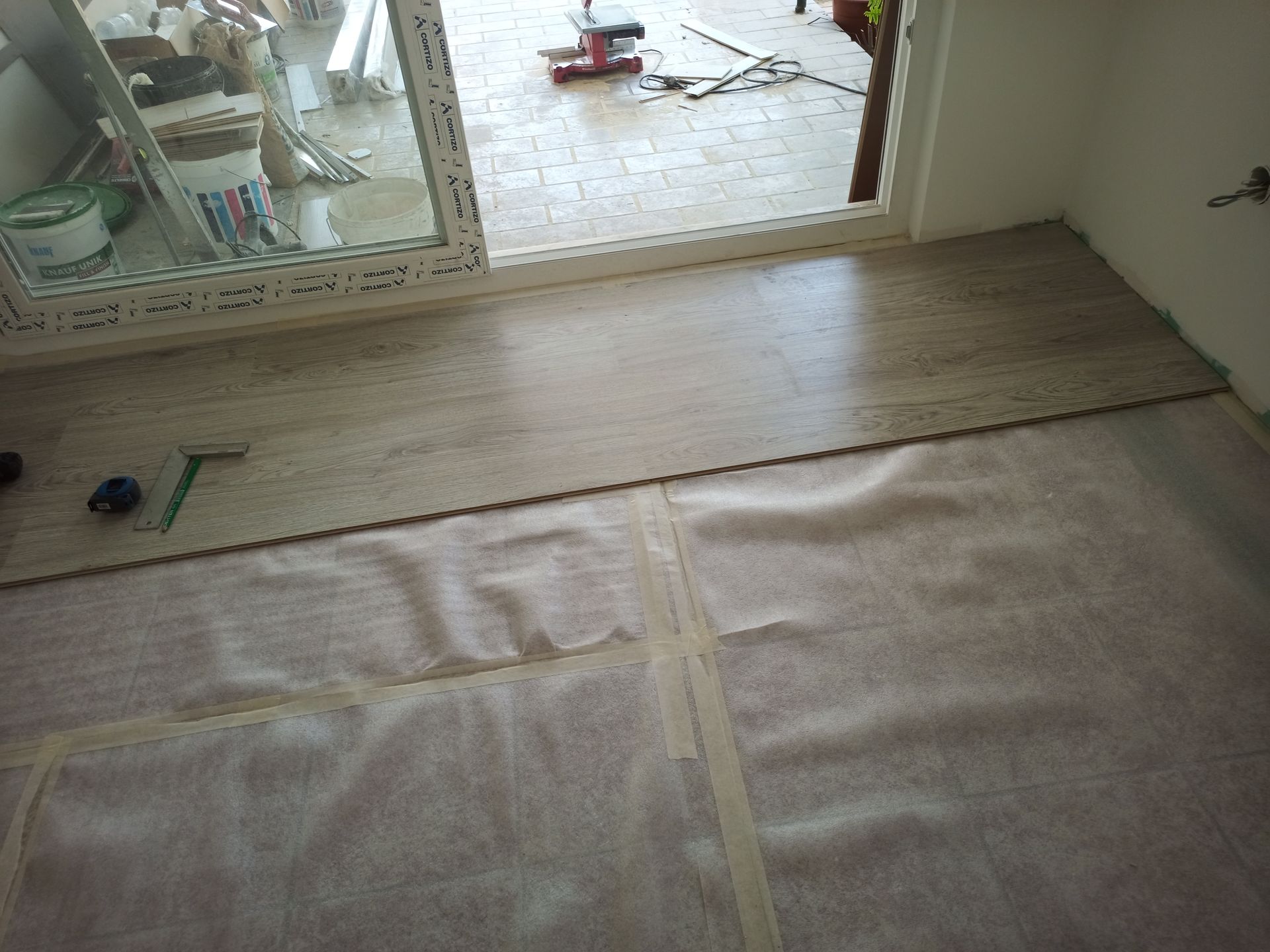 Instalação de piso: tábuas assentadas sobre manta acústica junto a uma porta de vidro deslizante. Materiais de construção visíveis.