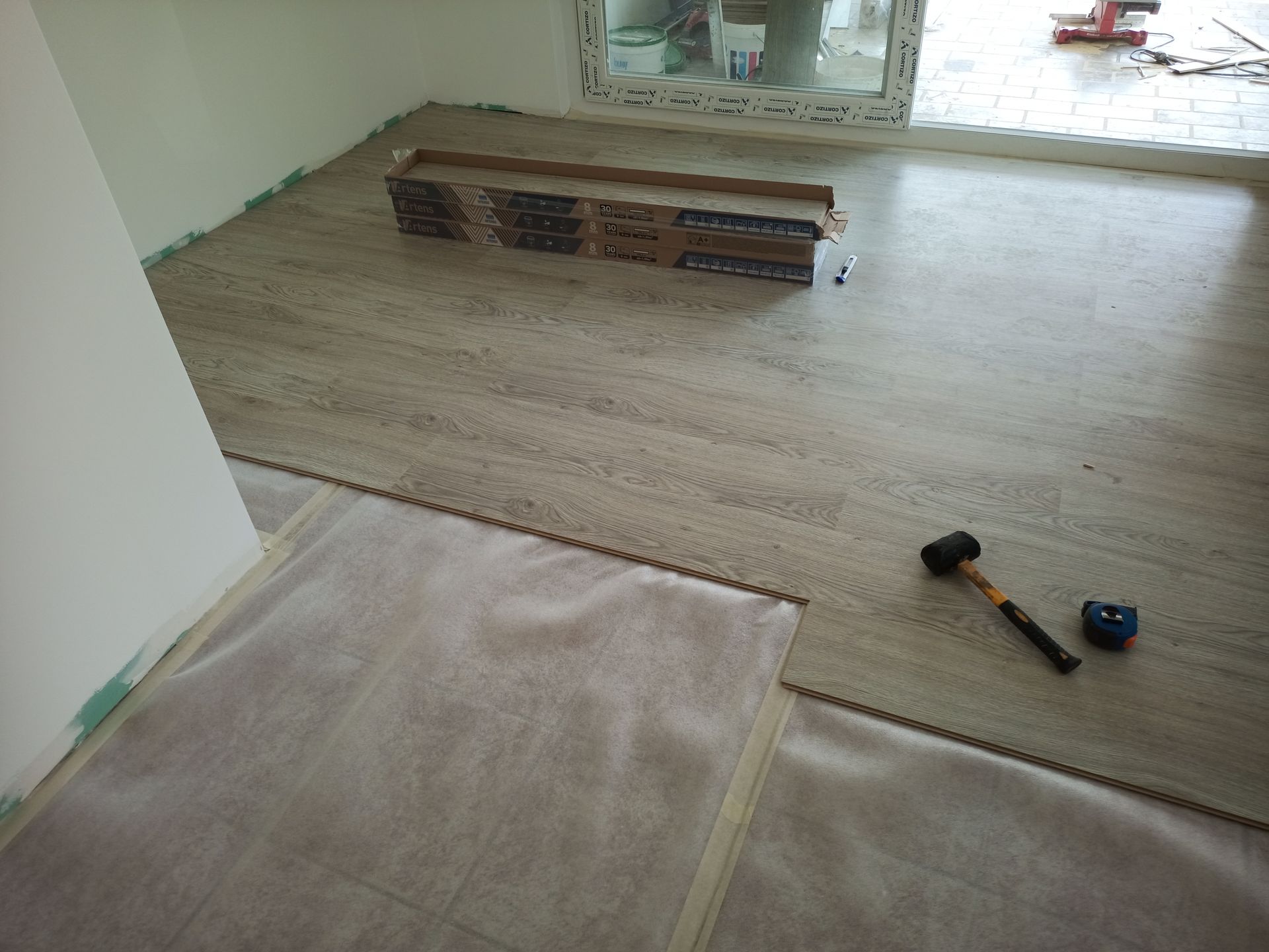 Instalação de piso laminado de cor clara em um cômodo com janela. O piso, um martelo e uma fita métrica estão visíveis.