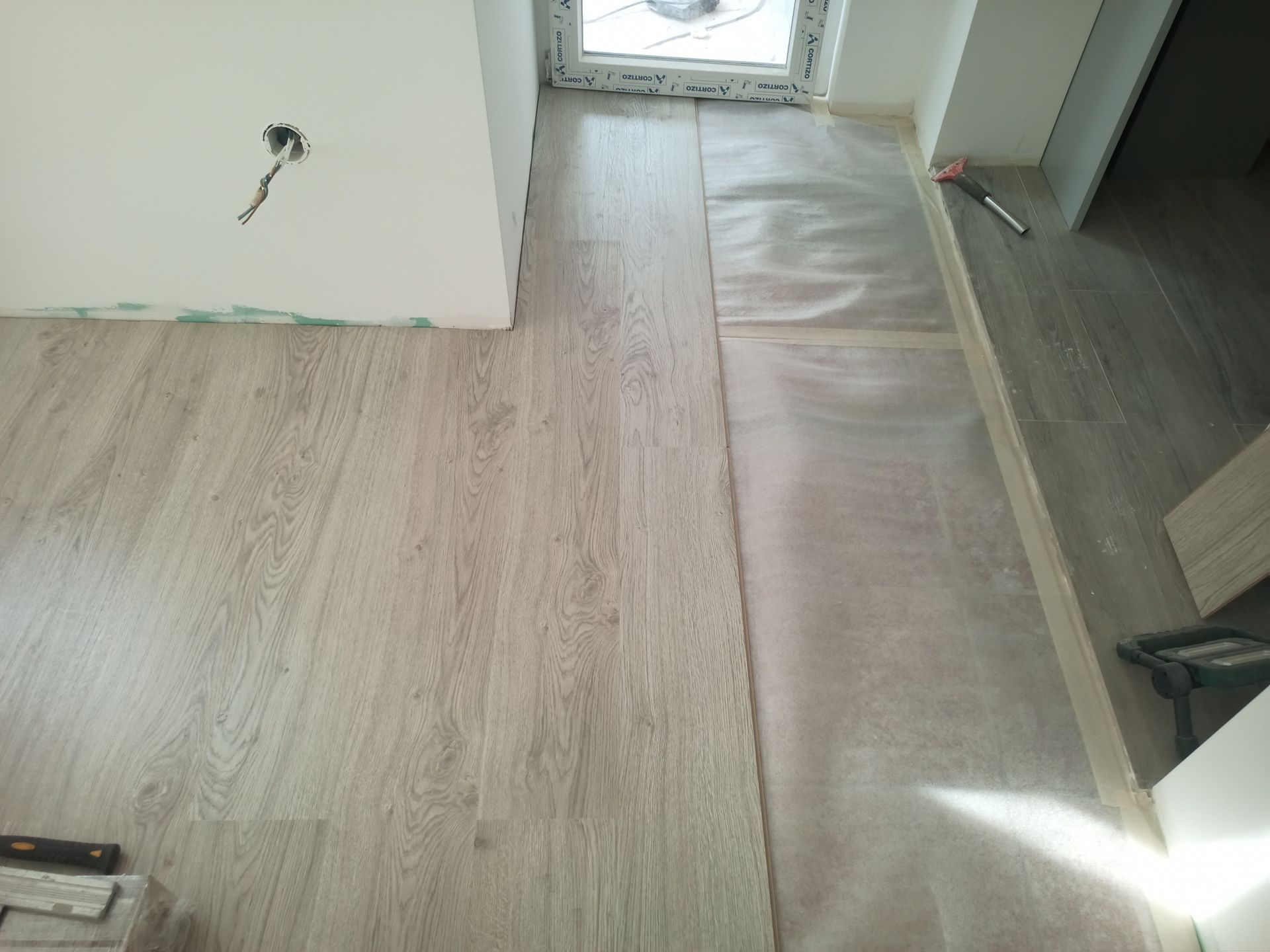 Instalação de piso laminado em andamento; tábuas colocadas junto a uma janela e porta.