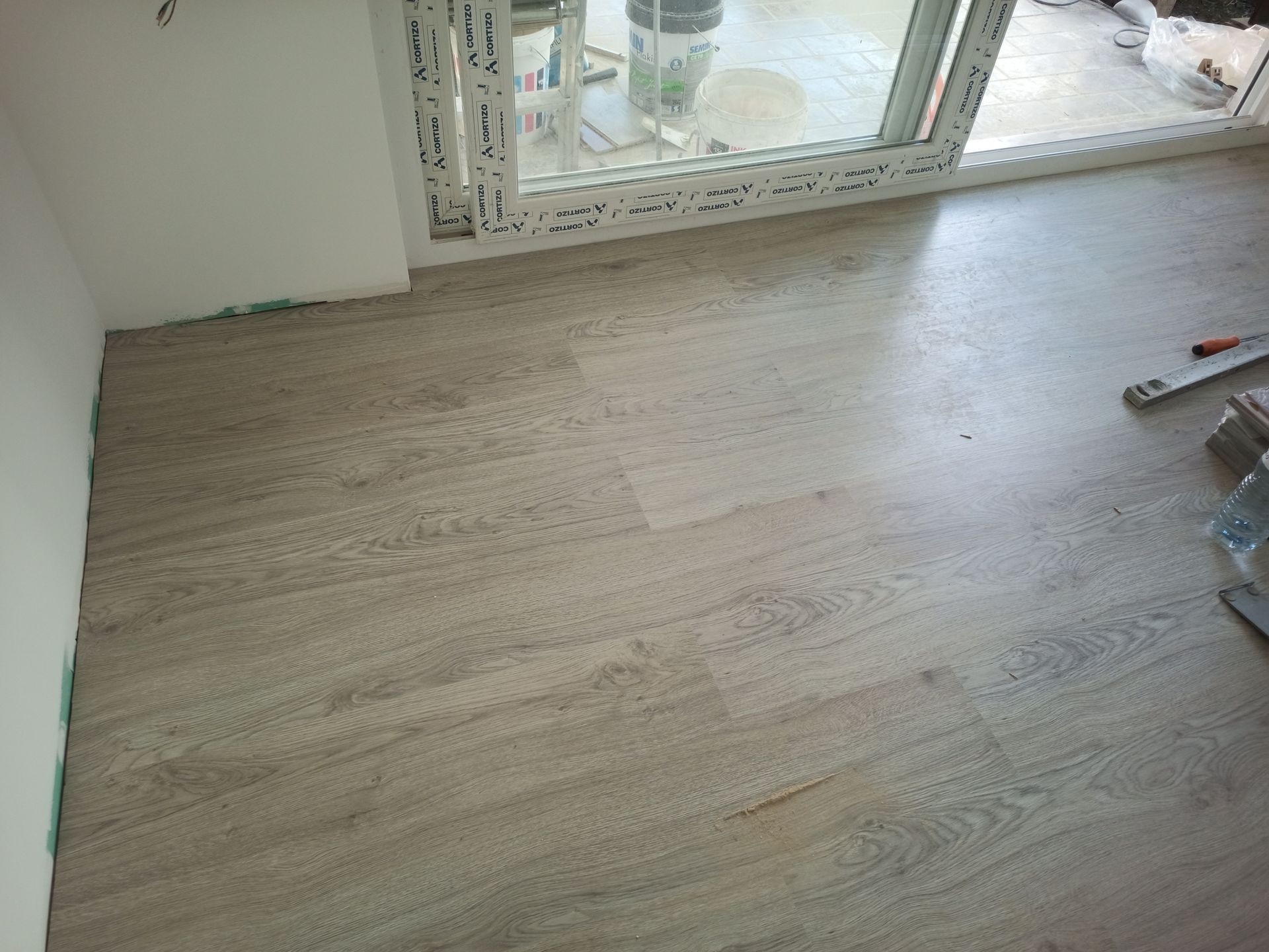 Piso com aparência de madeira clara instalado em um cômodo adjacente a uma parede branca e uma porta de vidro deslizante.