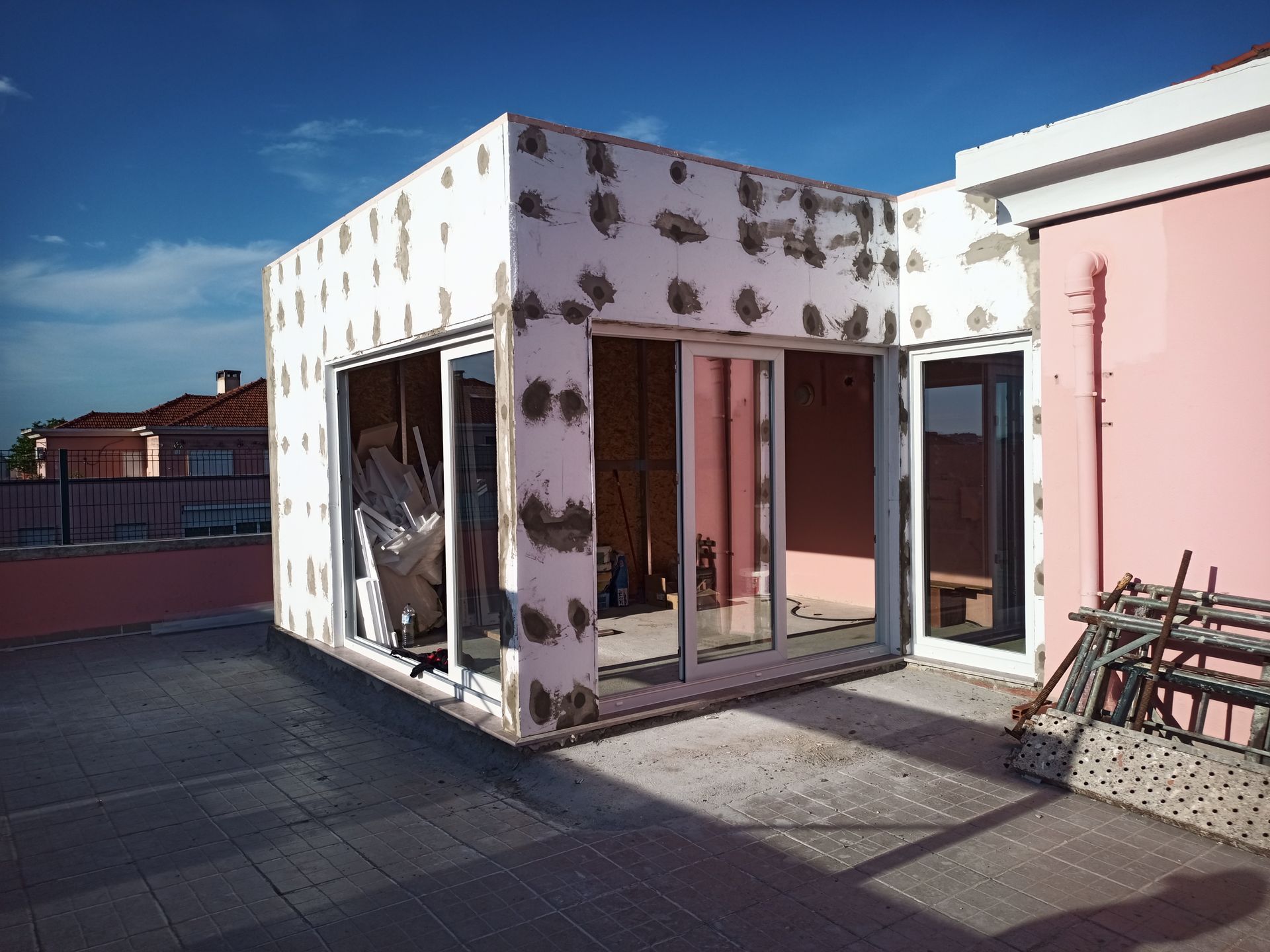 Construção de uma estrutura cúbica branca sobre um telhado plano. Isolamento aparente e portas de vidro. Prédio rosa ao fundo.