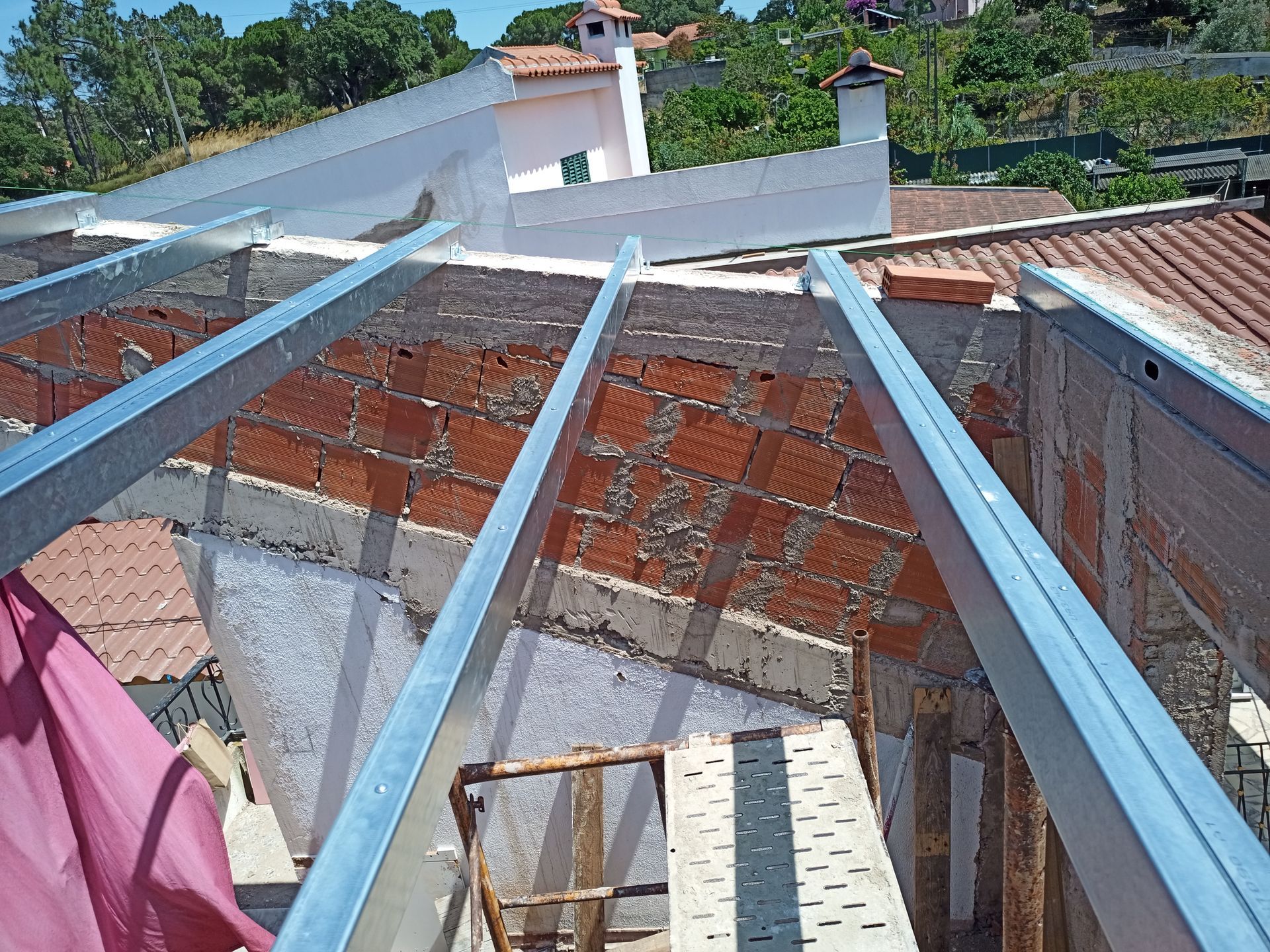 Vigas de metal dispostas transversalmente em uma parede de tijolos vermelhos, parte de um telhado em construção ao ar livre.