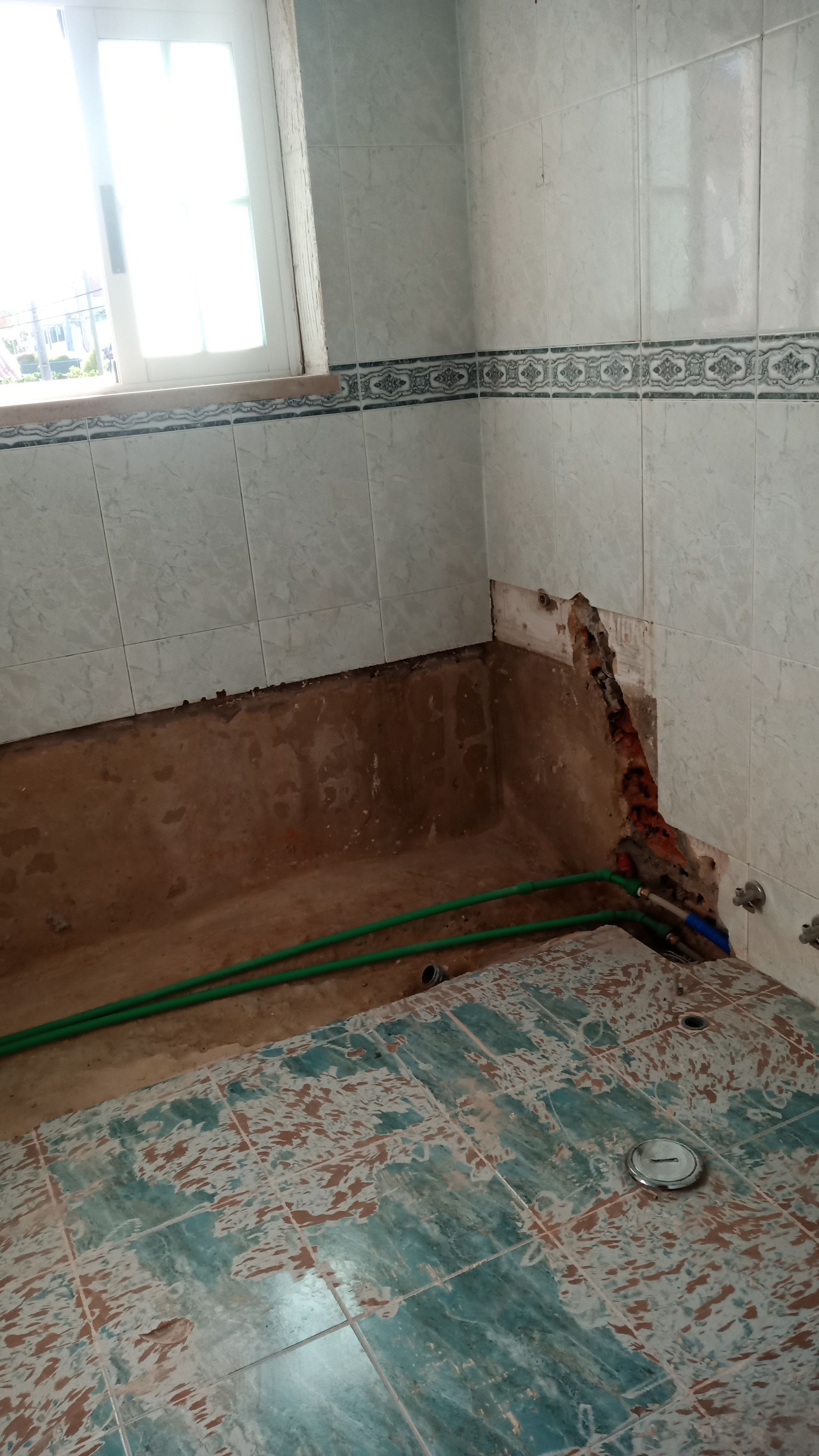 Reforma do banheiro em andamento, parede danificada, canos expostos, azulejos faltando, janela quebrada e entulho.