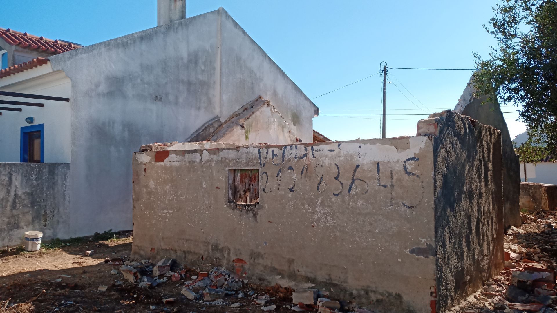 Vista exterior de um edifício parcialmente demolido ao lado de uma casa branca com moldura de porta azul sob um céu limpo.