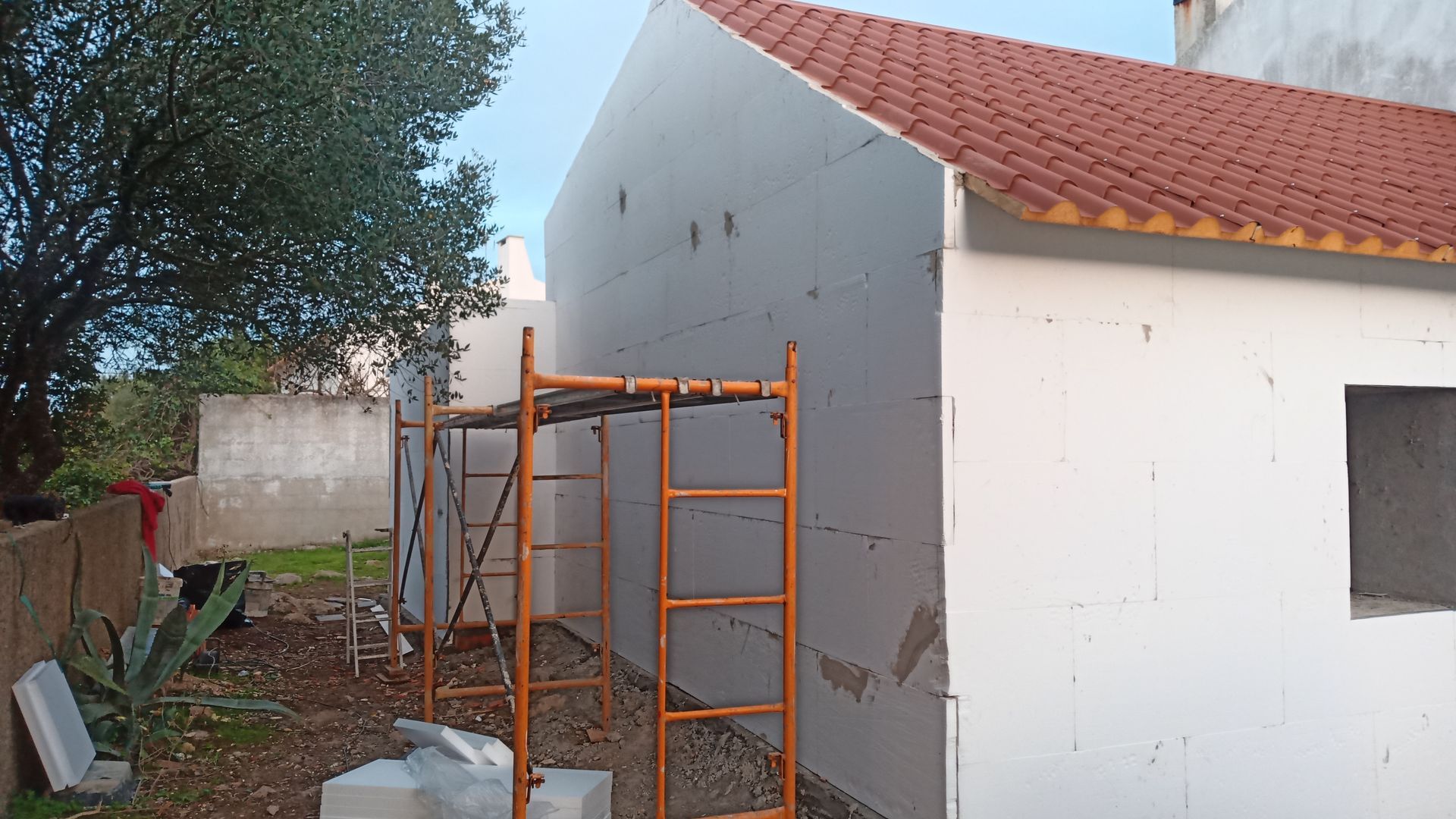 Canteiro de obras com andaimes encostado a um prédio branco com telhado de telhas vermelhas.